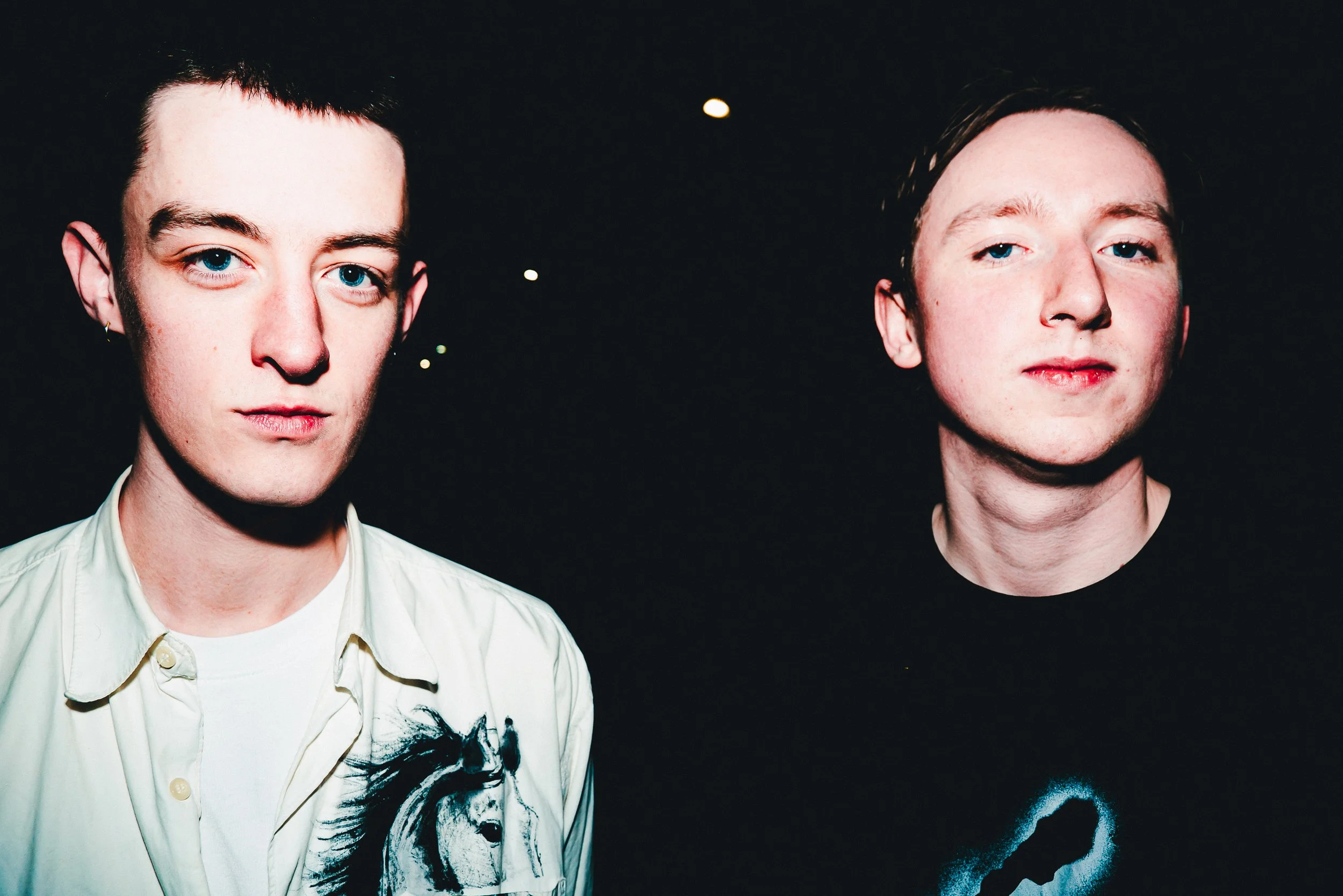 Close-up of two young men with light skin and blue eyes, standing side by side in a dark setting, facing the camera. The man on the left has dark hair, wears a light-colored shirt with a horse graphic, and an earring. The man on the right has short h