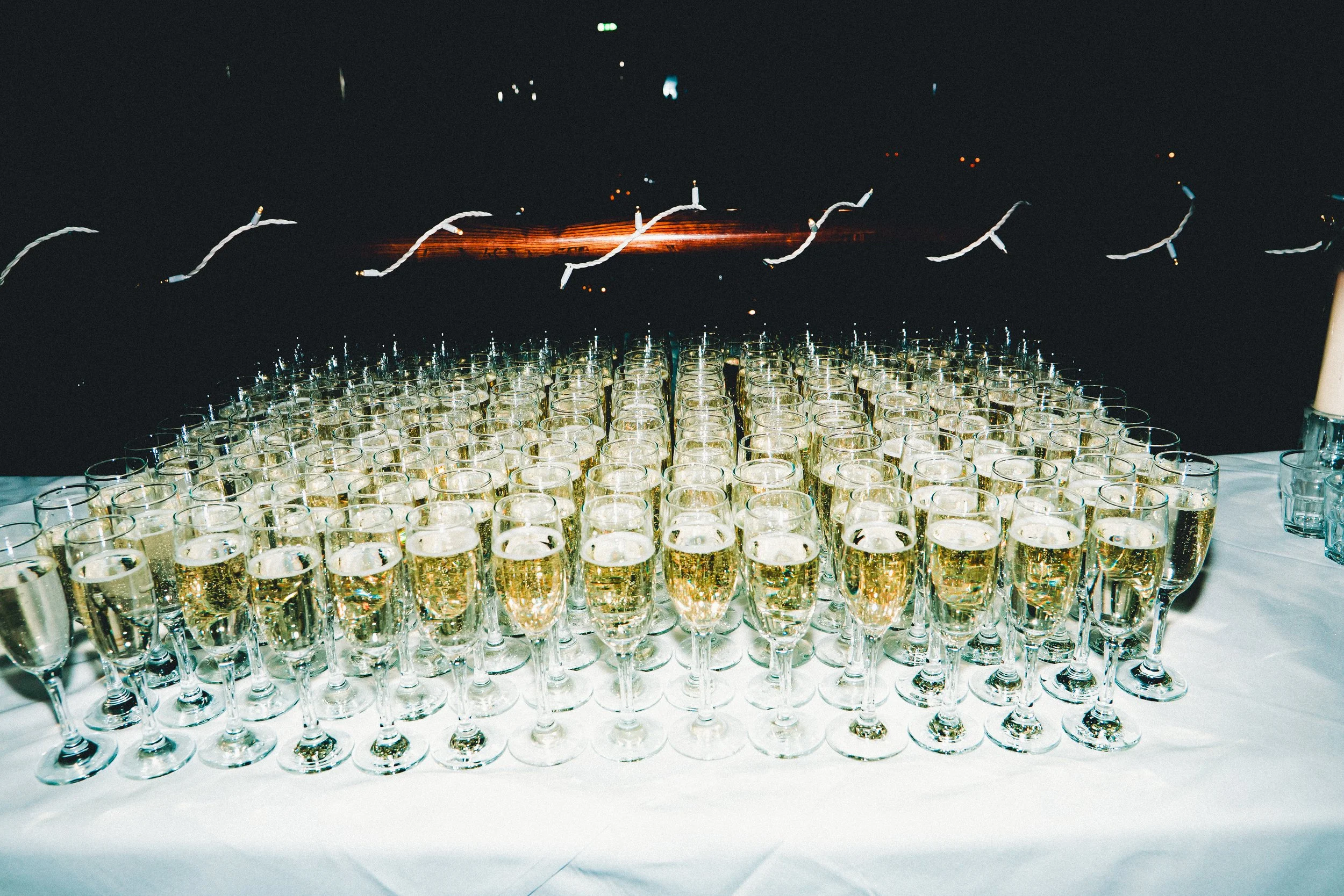 A large number of champagne glasses filled with sparkling wine arranged on a white tablecloth. In the background, a dark area with some blurred lights and candles, creating a festive atmosphere.