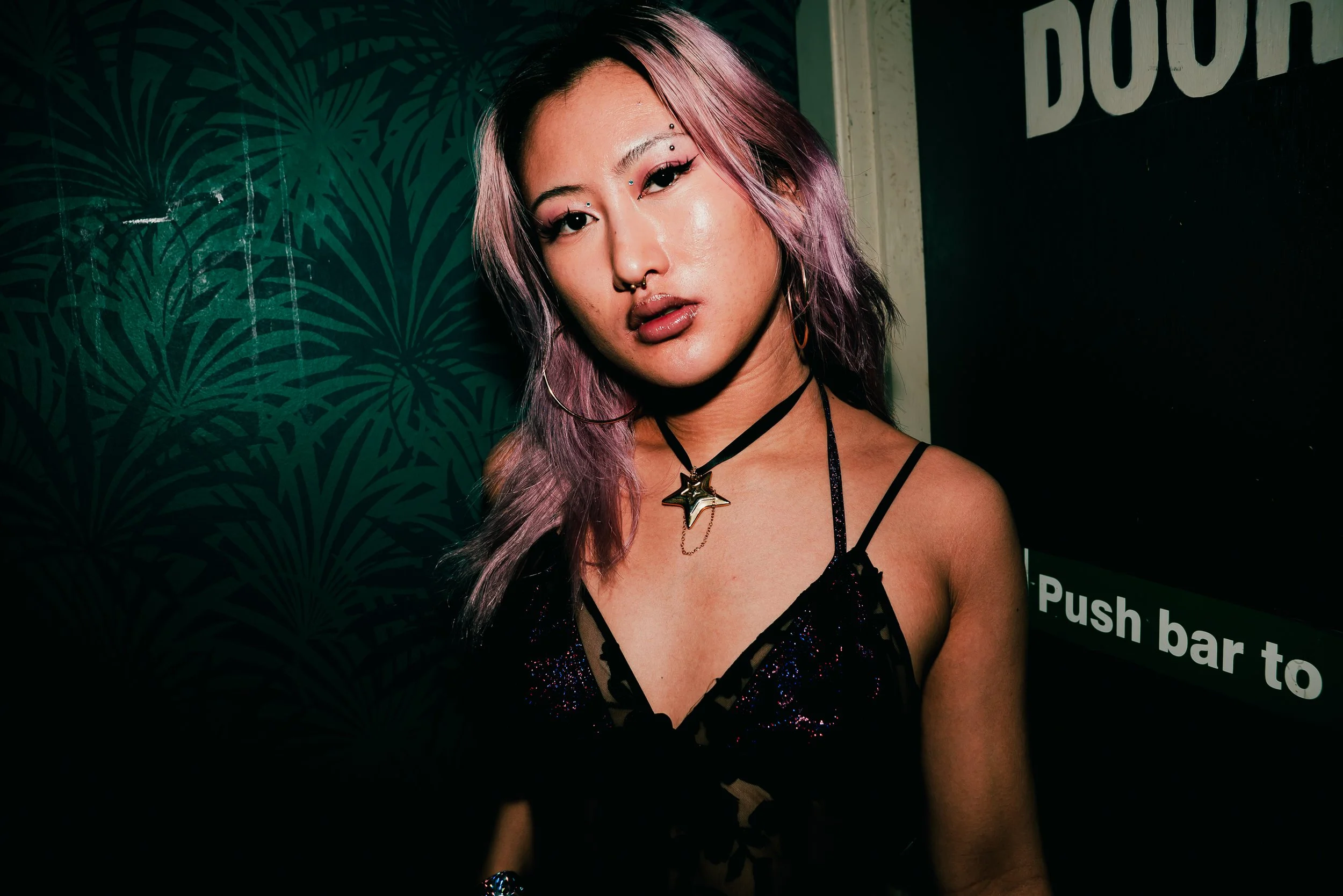 A woman with pinkish-purple hair styled in loose waves, wearing makeup including winged eyeliner, blush, and glossy lipstick. She has facial piercings including a septum ring and piercings above her eyebrows. She is dressed in a black, strappy, spark