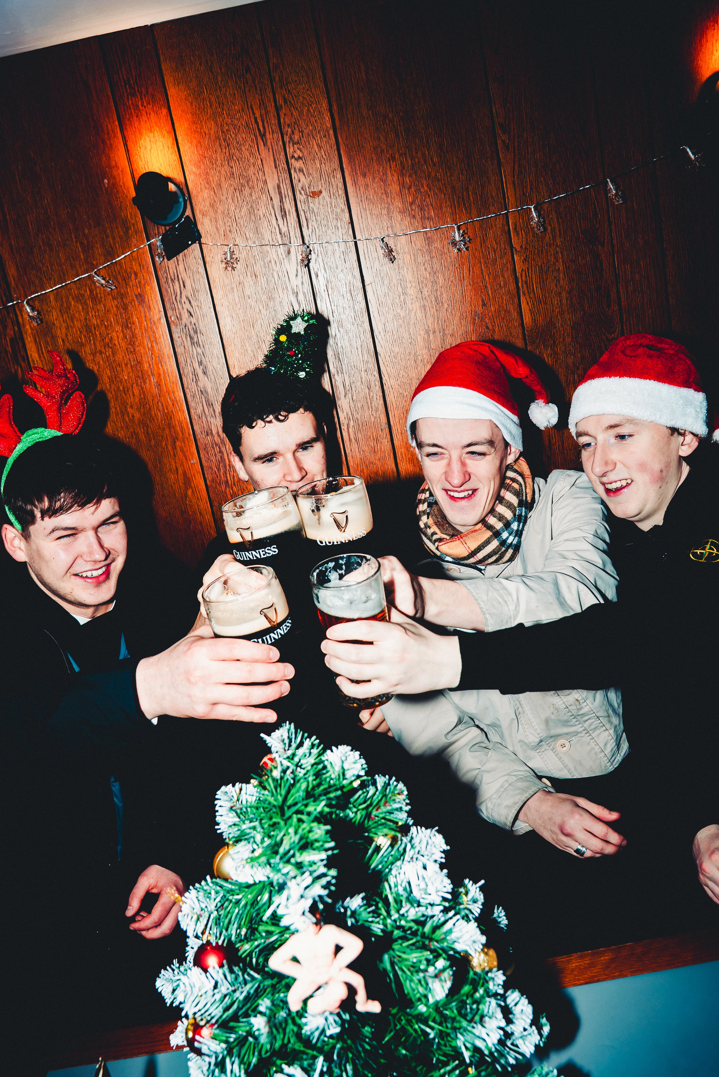 Group of friends celebrating Christmas, wearing Santa hats, raising glasses of beer, near a decorated Christmas tree with a wooden wall background.