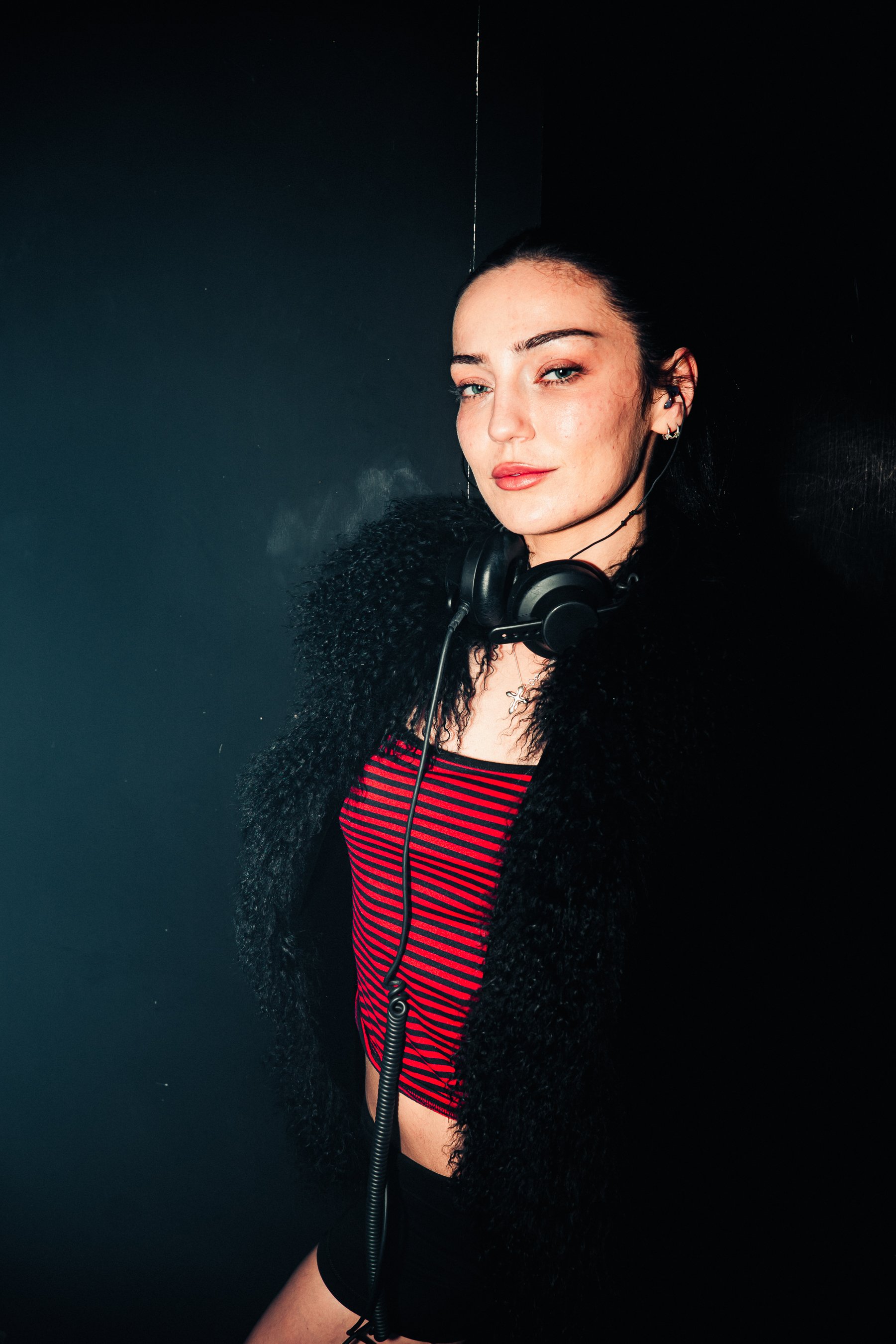 A woman with black curly hair, light skin, and blue eyes wearing headphones, a black fuzzy jacket, and a red and black striped shirt, standing against a dark background.
