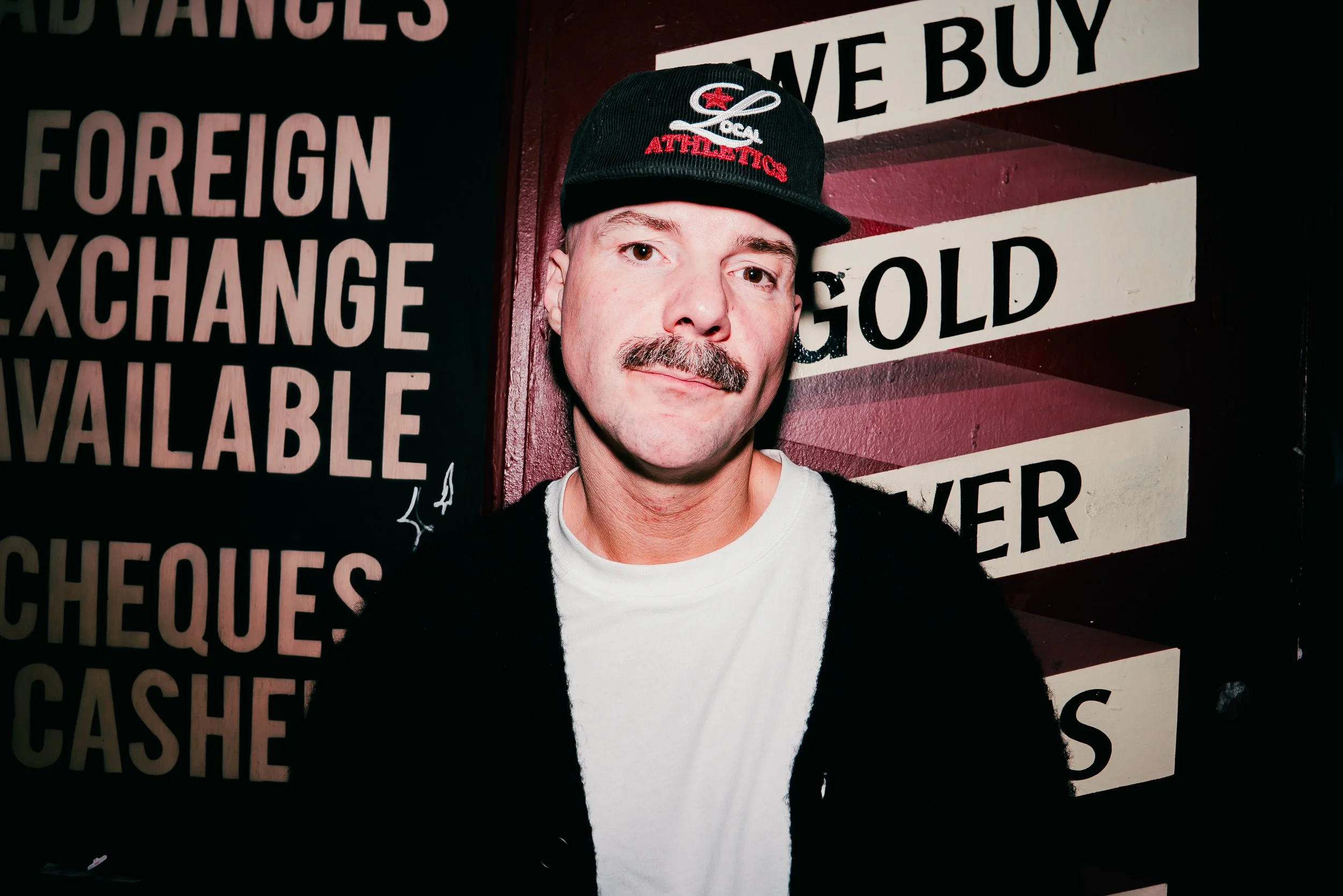 A man with a mustache and wearing a black cap with red and white embroidery, including the word 'ATHLETICS,' stands in front of a sign with black and white text on a dark red background. The sign reads 'ID VANGLES,' 'FOREIGN,' 'EXCHANGE,' 'AVAILABLE,