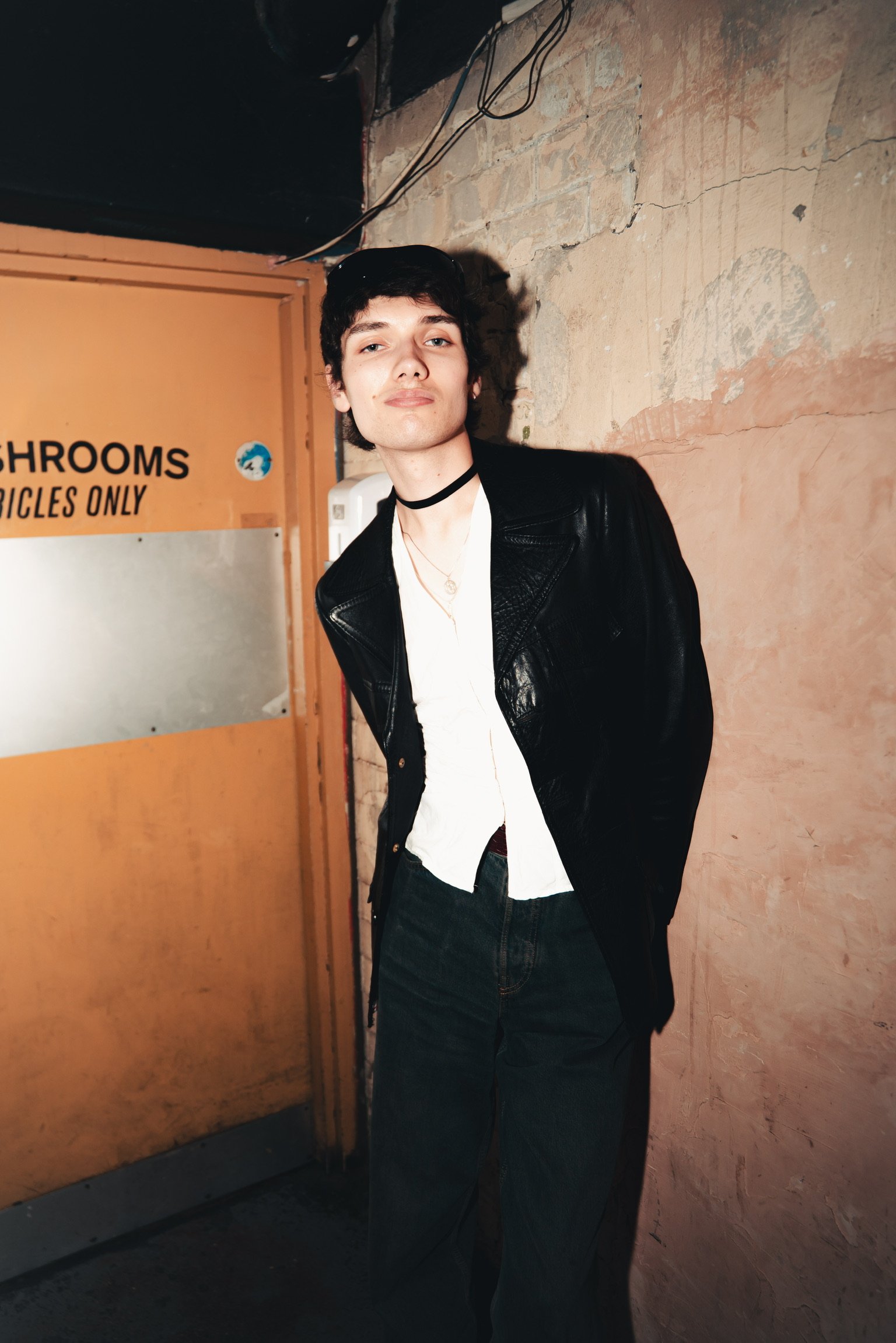 A young man with dark hair wearing a black leather jacket, white shirt, and a black choker necklace standing in front of a brick wall and a yellow-orange door with the words "MUSHROOMS BICYCLES ONLY" on it, in an indoor setting.