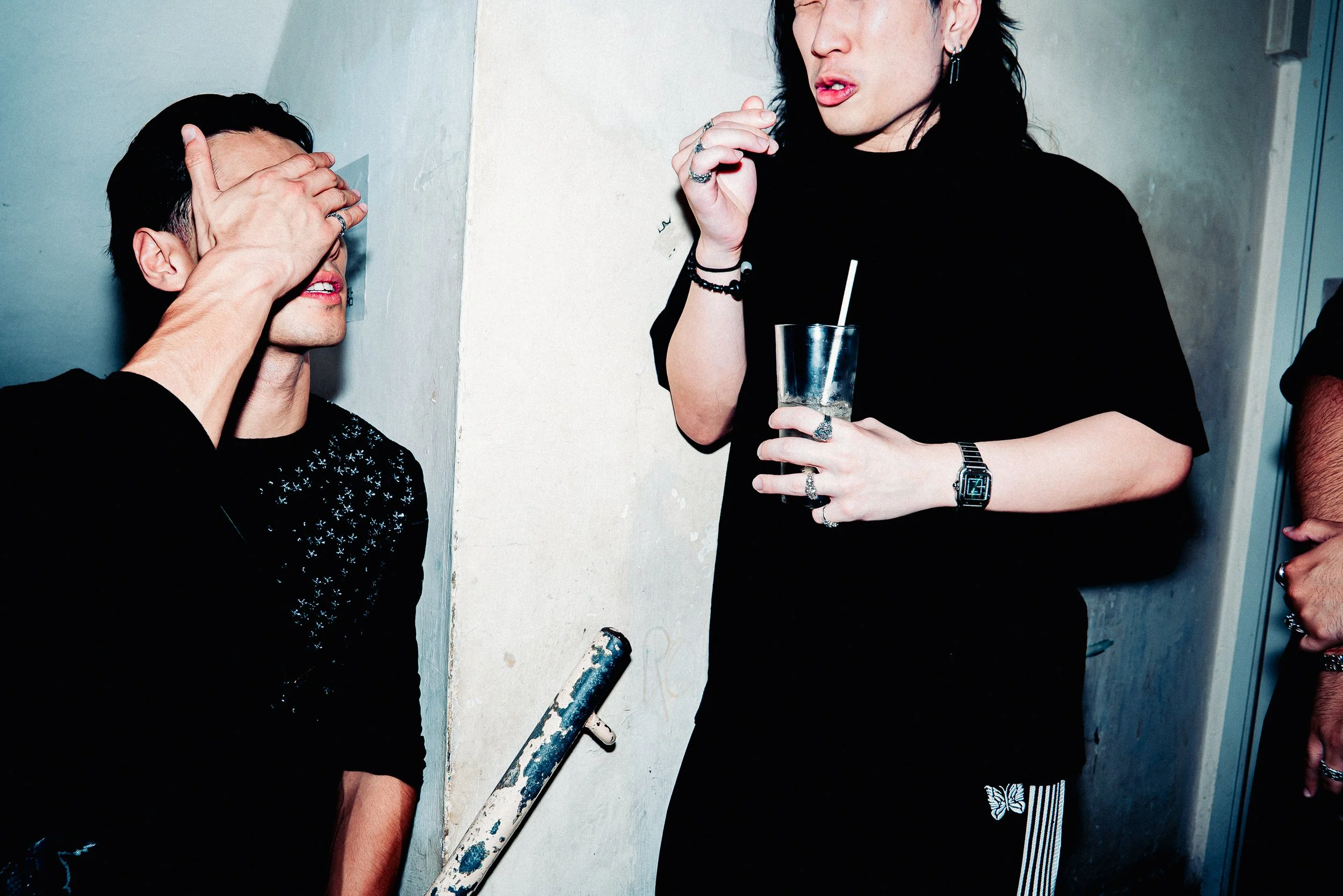 Two women are in conversation in an indoor setting. One woman has short dark hair and covers her face with her hand, while the other woman has long dark hair, jewelry, and holds a glass with a straw.