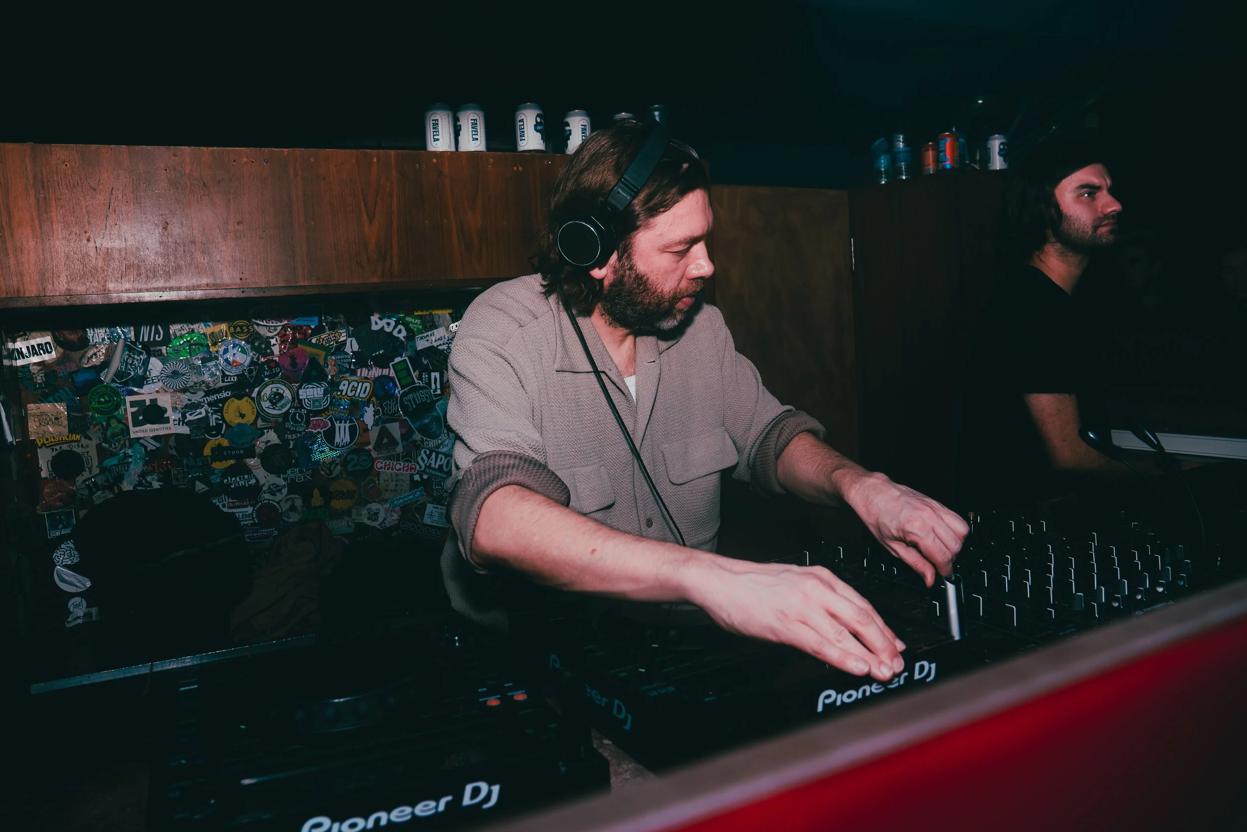 DJ with headphones operating turntables at a music event, with a sticker-covered wall behind him.