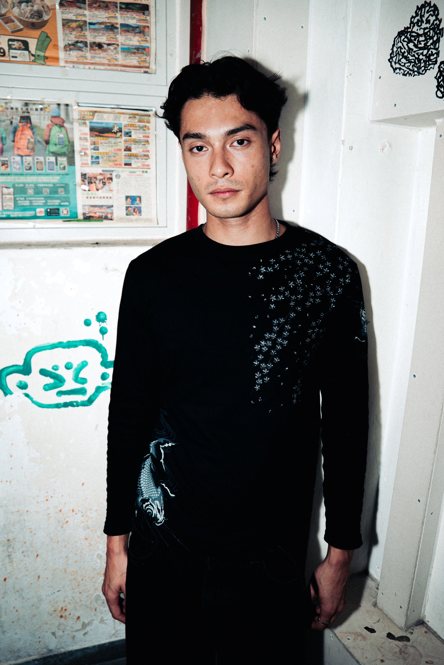 A young man with dark hair, wearing a black shirt with a pattern, standing in front of a white wall with posters and graffiti.