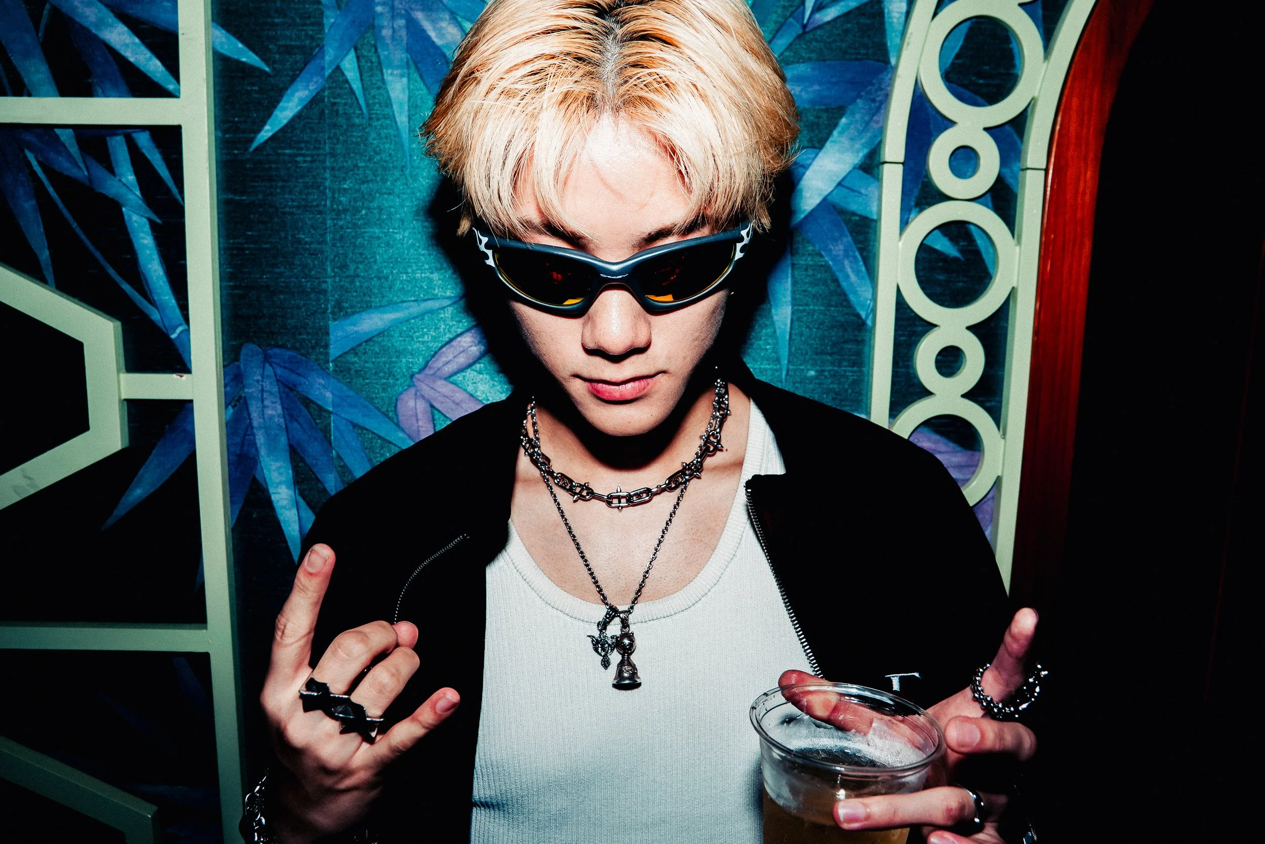A young person with light blonde hair wearing sunglasses and layered necklaces, holding a clear cup with a dark beverage, making a peace sign with both hands. They are dressed in a black jacket over a white shirt, standing in front of a colorful, abs