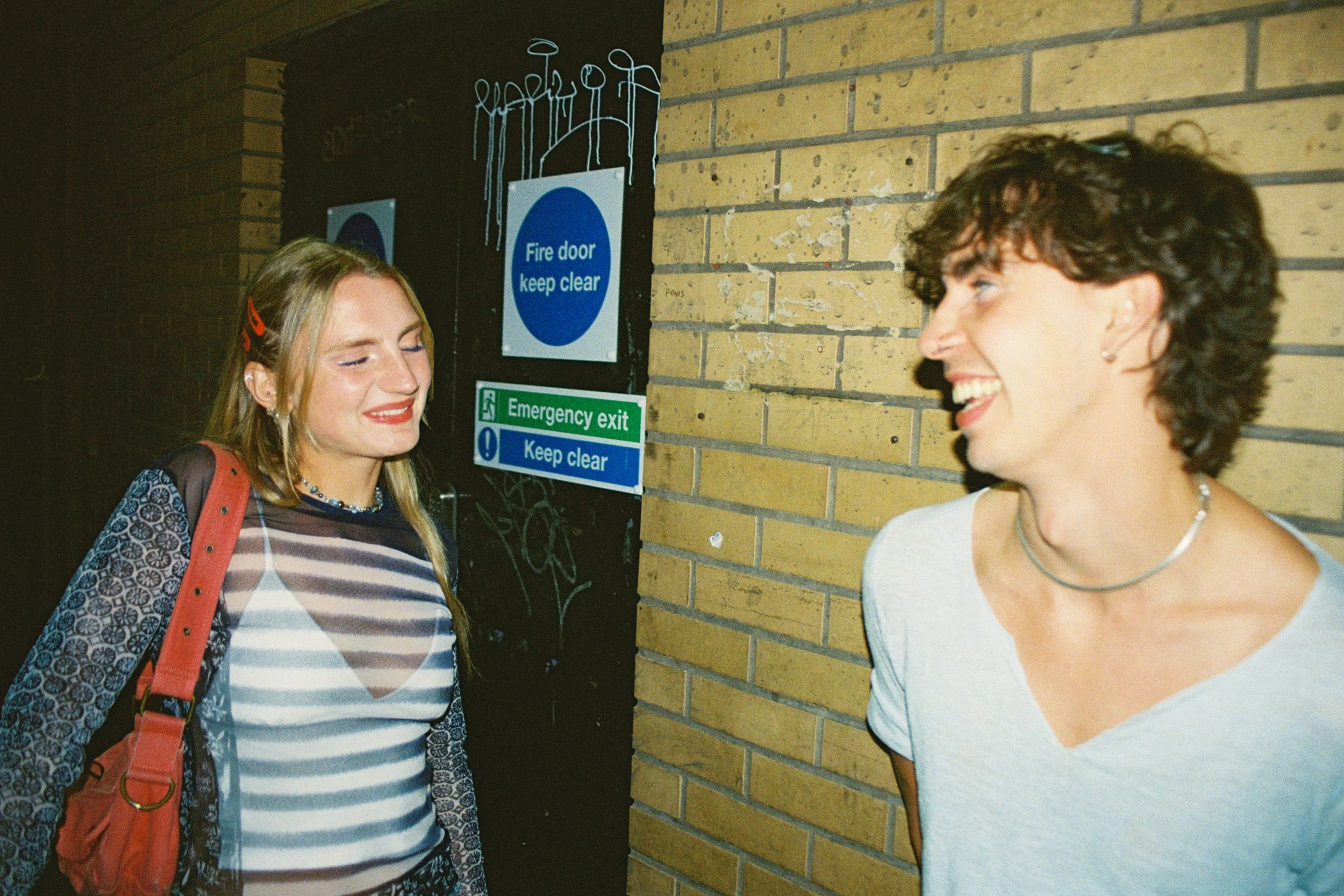 Two women smiling and talking near a brick wall, with signs about fire safety and emergency exit behind them.