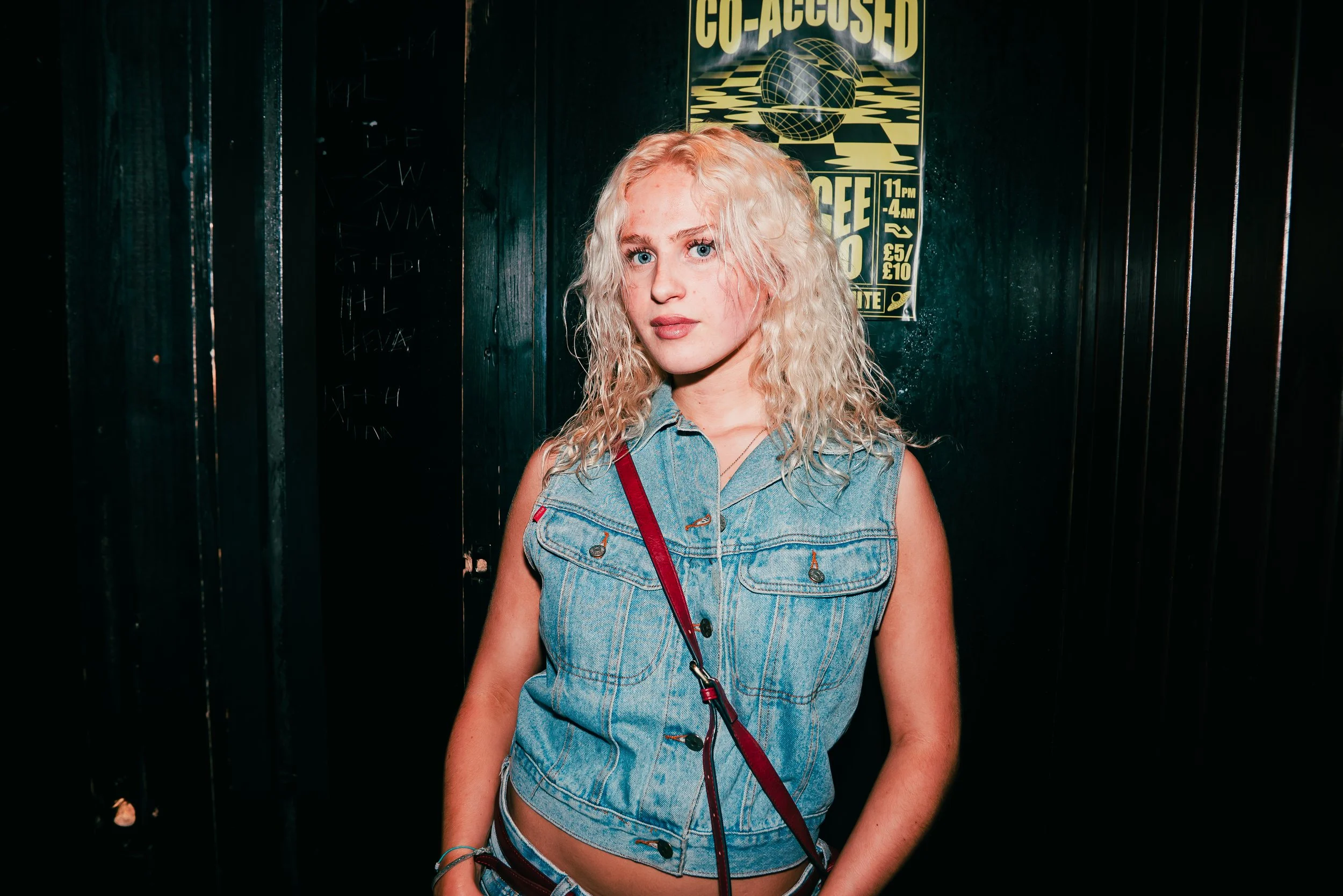 A young woman with curly blonde hair, light skin, and blue eyes standing in front of a dark wall with a yellow and black poster that reads 'CO-ASSURED'. She is wearing a sleeveless denim top and has a red crossbody bag.