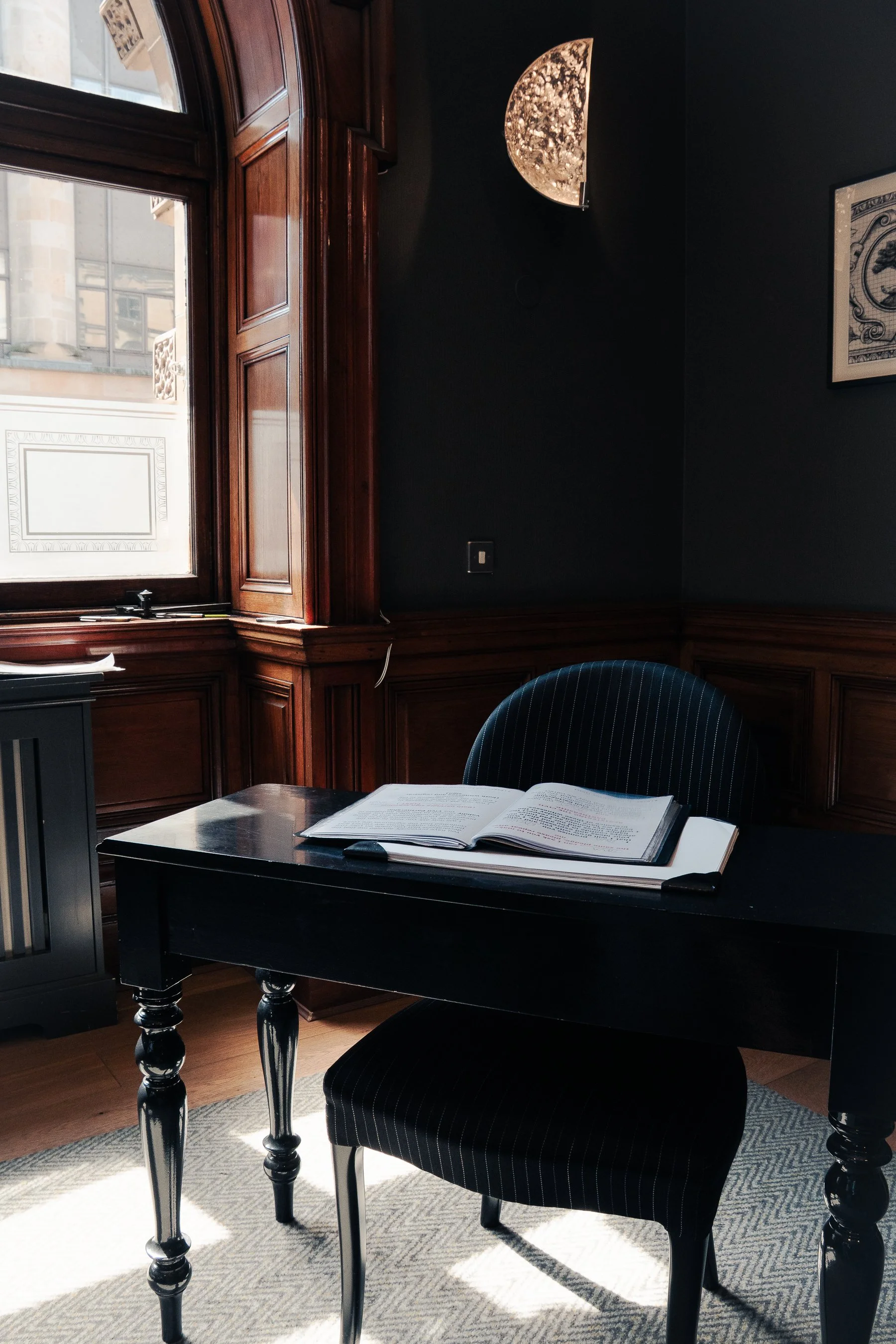 A study or office space with a black wooden desk, an open book, a dark upholstered chair, wood-paneled walls, and a window with wooden trim. There is framed artwork on the wall and a textured window covering that resembles a moon.