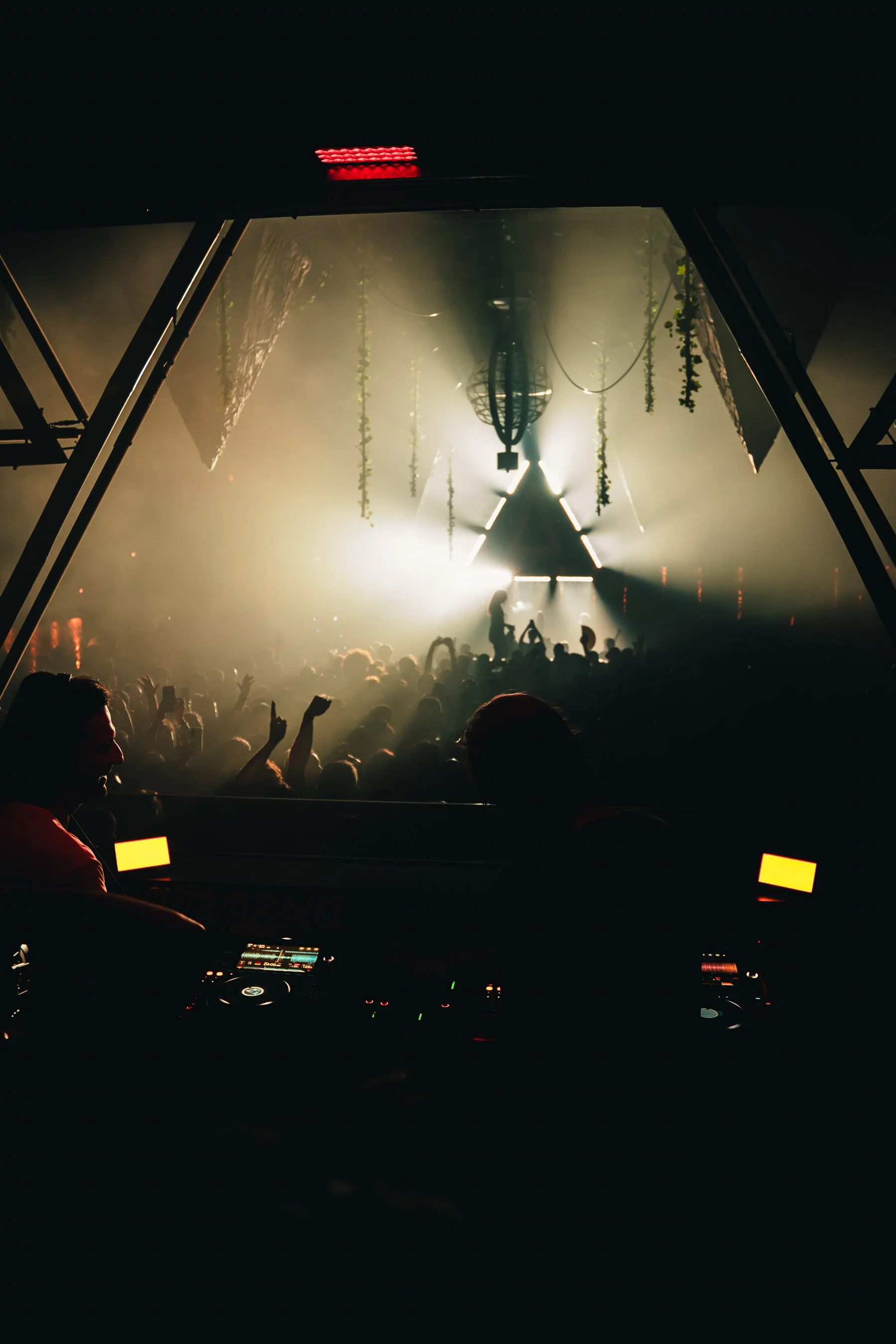 Inside a nightclub or concert venue with fog and bright lights, a DJ is playing at a booth in the foreground, and a large crowd is dancing and enjoying the event, with some holding up their hands.