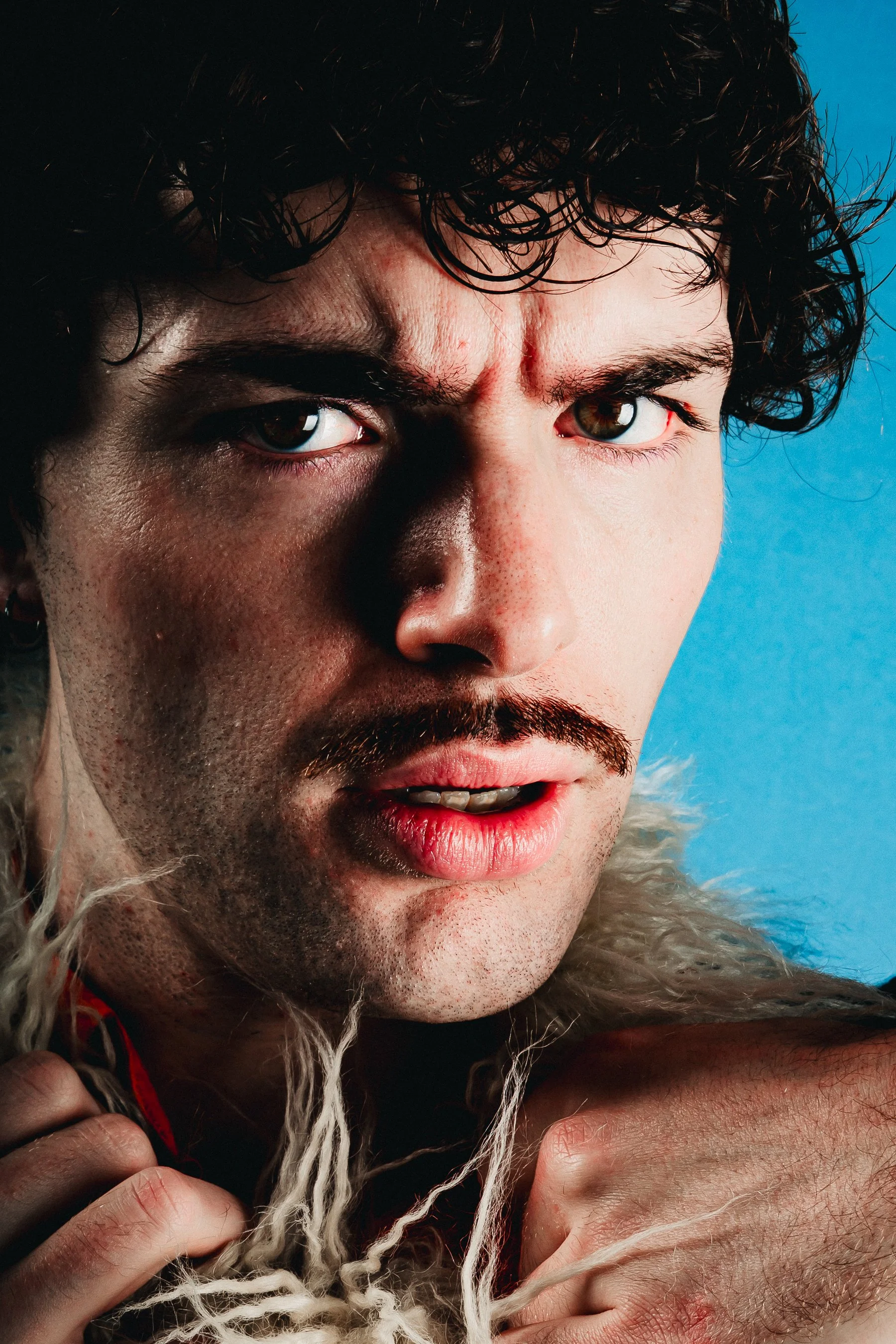 Close-up of a man with curly hair, brown eyes, facial hair, and wearing a thick, fur-lined coat against a blue background.