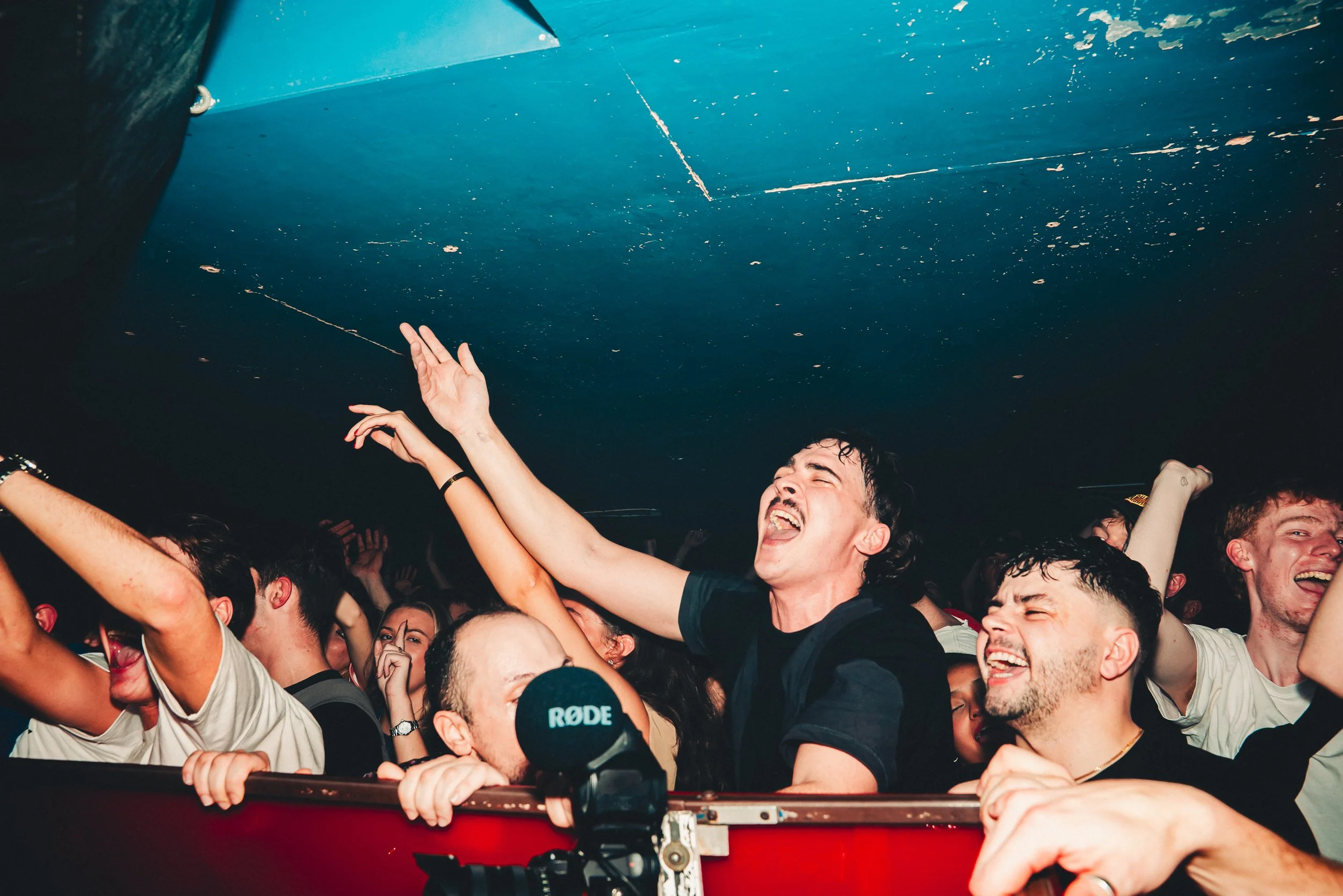 People dancing and enjoying a live concert or event, with a musician in the foreground wearing headphones and raising his hand, surrounded by a crowd in a dark indoor venue.