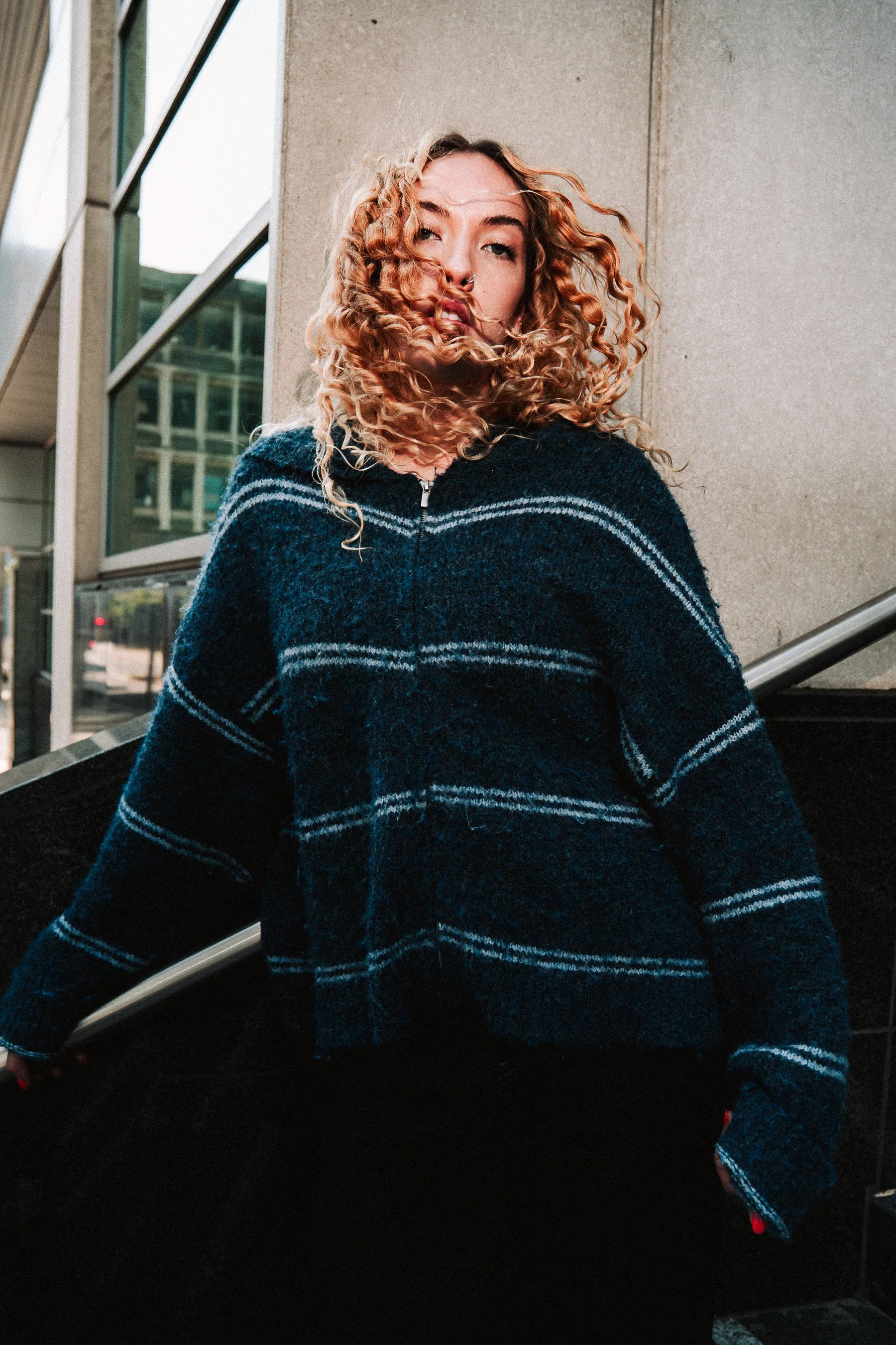 A woman with curly red hair and a septum piercing, wearing a dark blue plaid sweater, sits on a staircase outside a modern building with large glass windows and concrete walls.