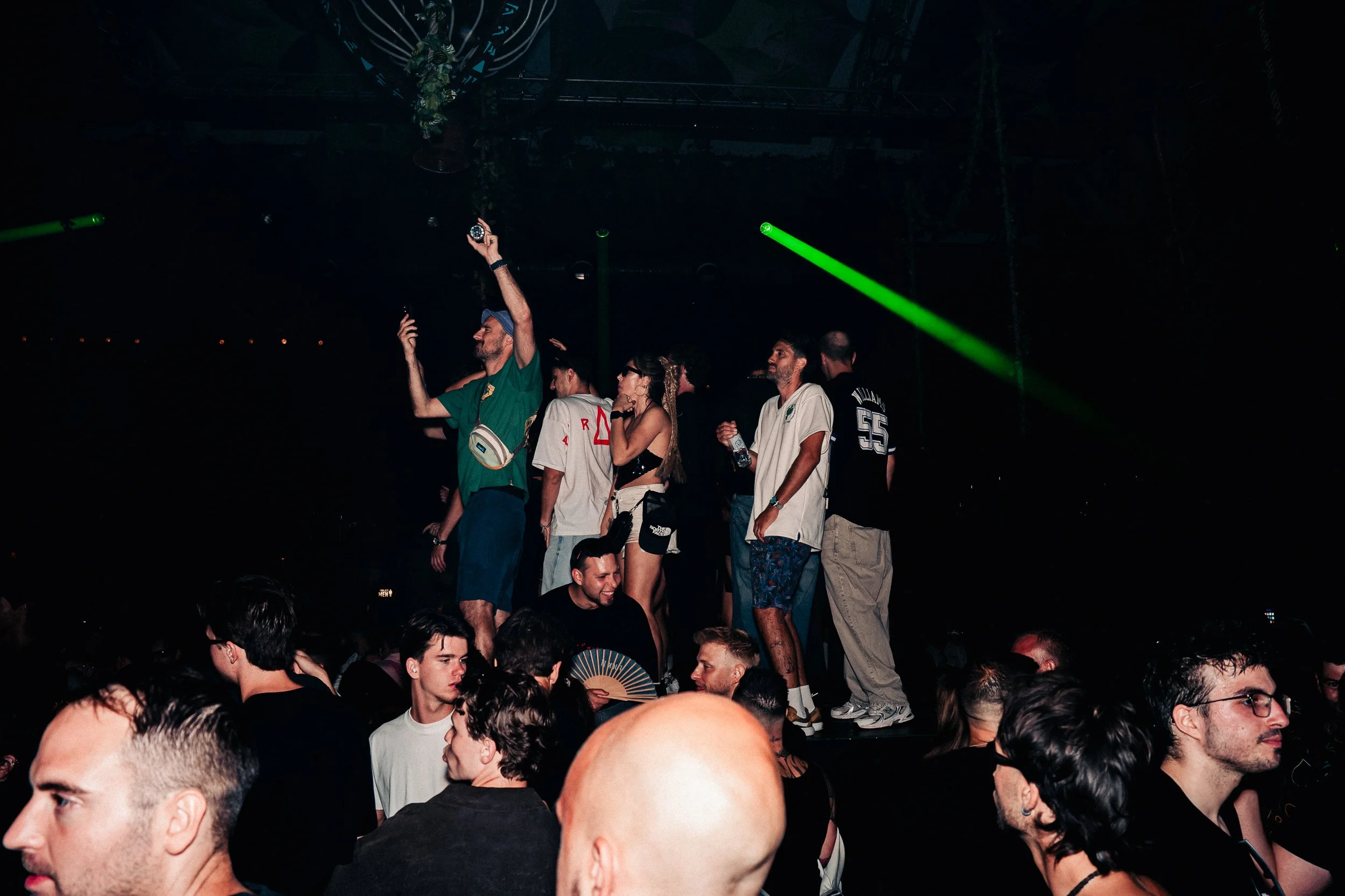 People standing and sitting in a crowded dark indoor venue with green laser lights overhead, some talking and enjoying the event.