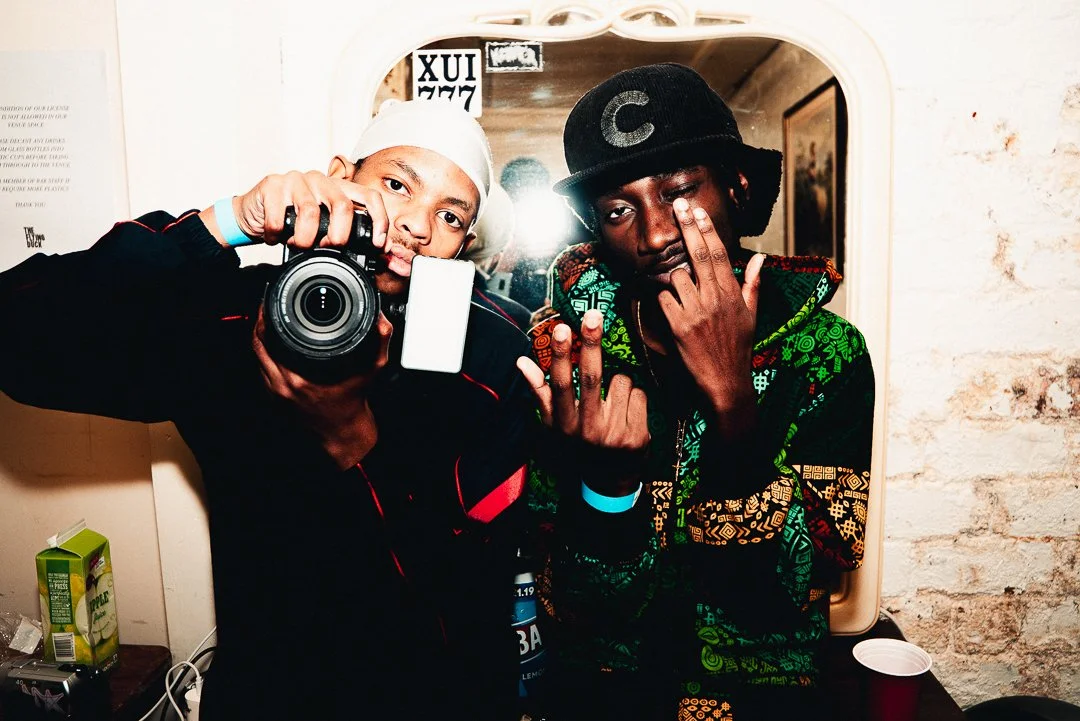 Two young men taking a mirror selfie. One is holding a camera, wearing a white cap, and the other is making a gesture with his hand, wearing a black cap and a colorful hoodie.