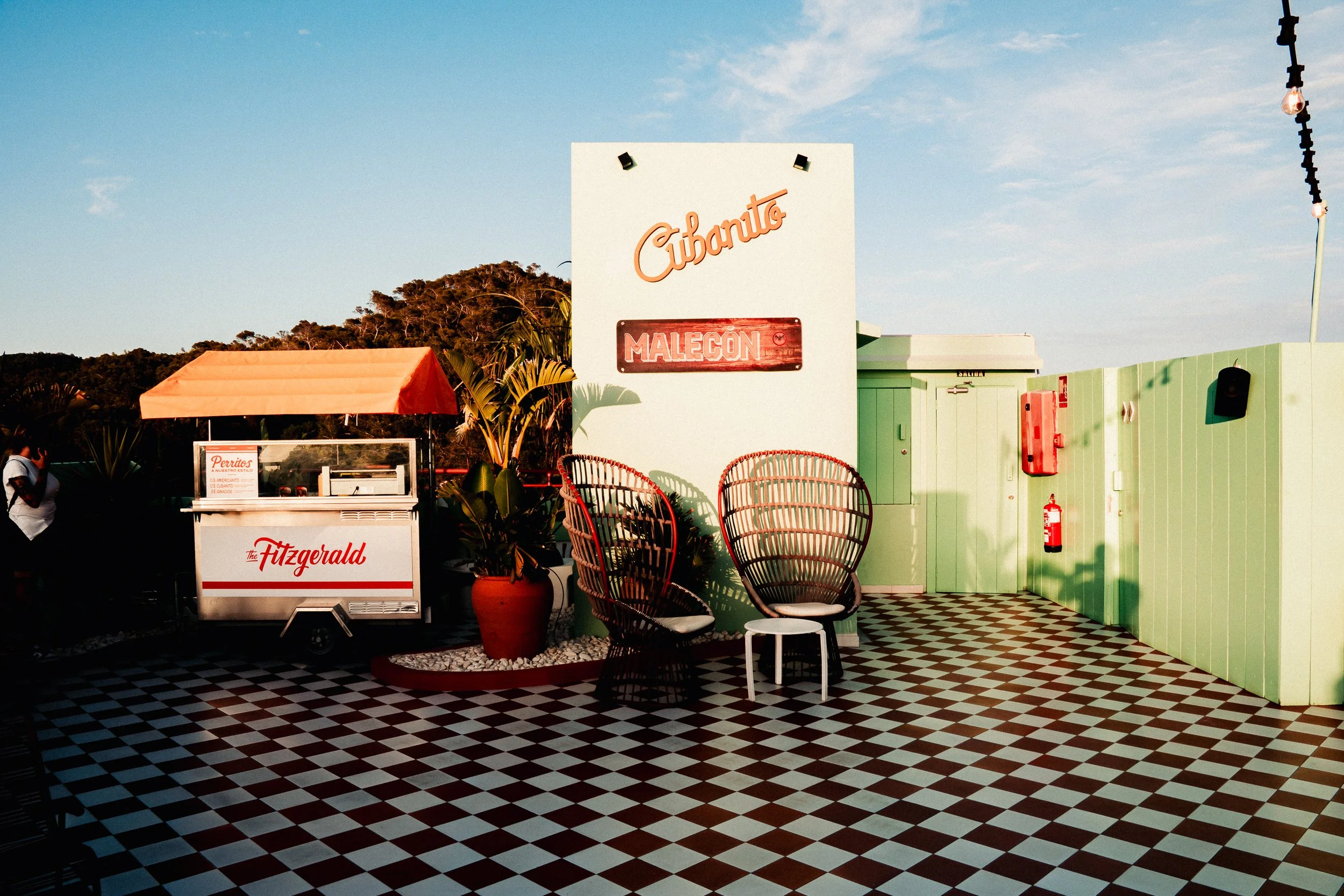 Outdoor seating area with checkered black and white floor, a large white sign with orange script text 'Cibanto' and a smaller red sign 'Malecon', a green wall with fire extinguisher and a black speaker, red chairs, a potted plant, and a food cart wit