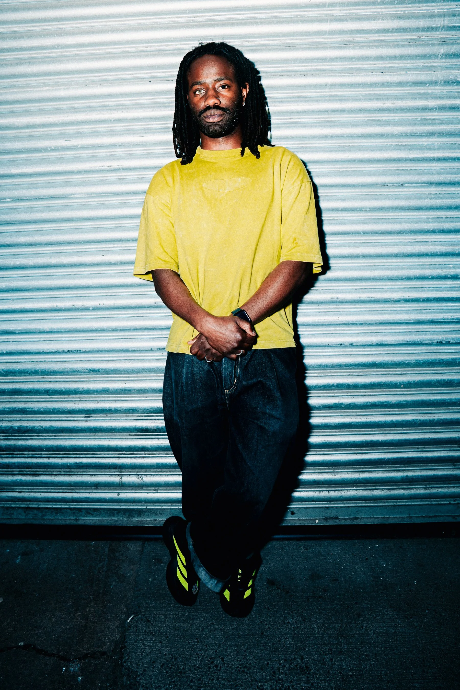 A man with dreadlocks and a beard wearing a yellow T-shirt, black pants, and black and yellow sneakers standing against a metallic roll-up door, with one leg crossed over the other and arms folded.