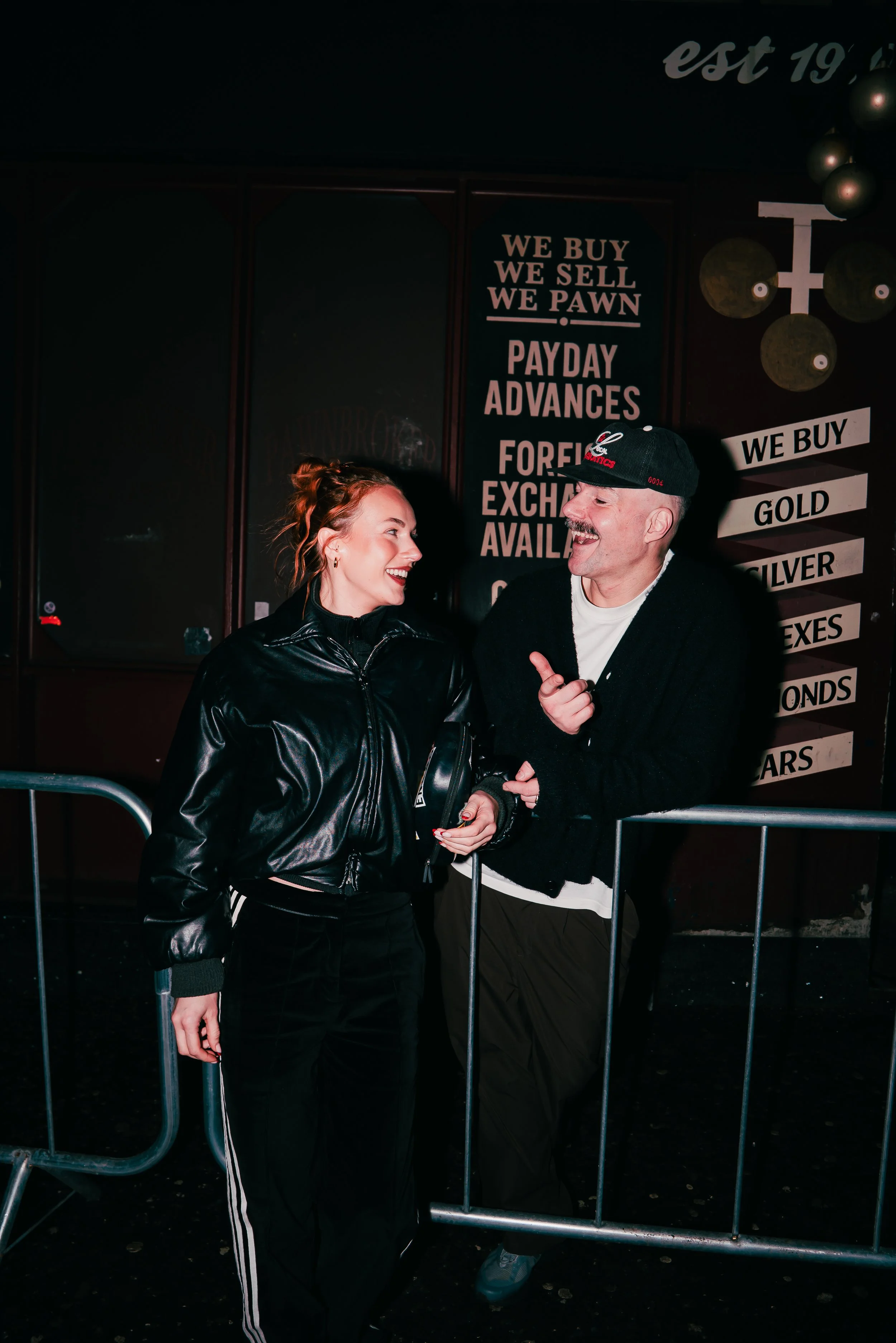 A man and a woman are smiling and talking to each other behind a metal crowd barrier at night. The woman has red hair styled in a bun, is wearing a shiny black double-breasted jacket, and black pants with white stripes. The man has a mustache, is wea