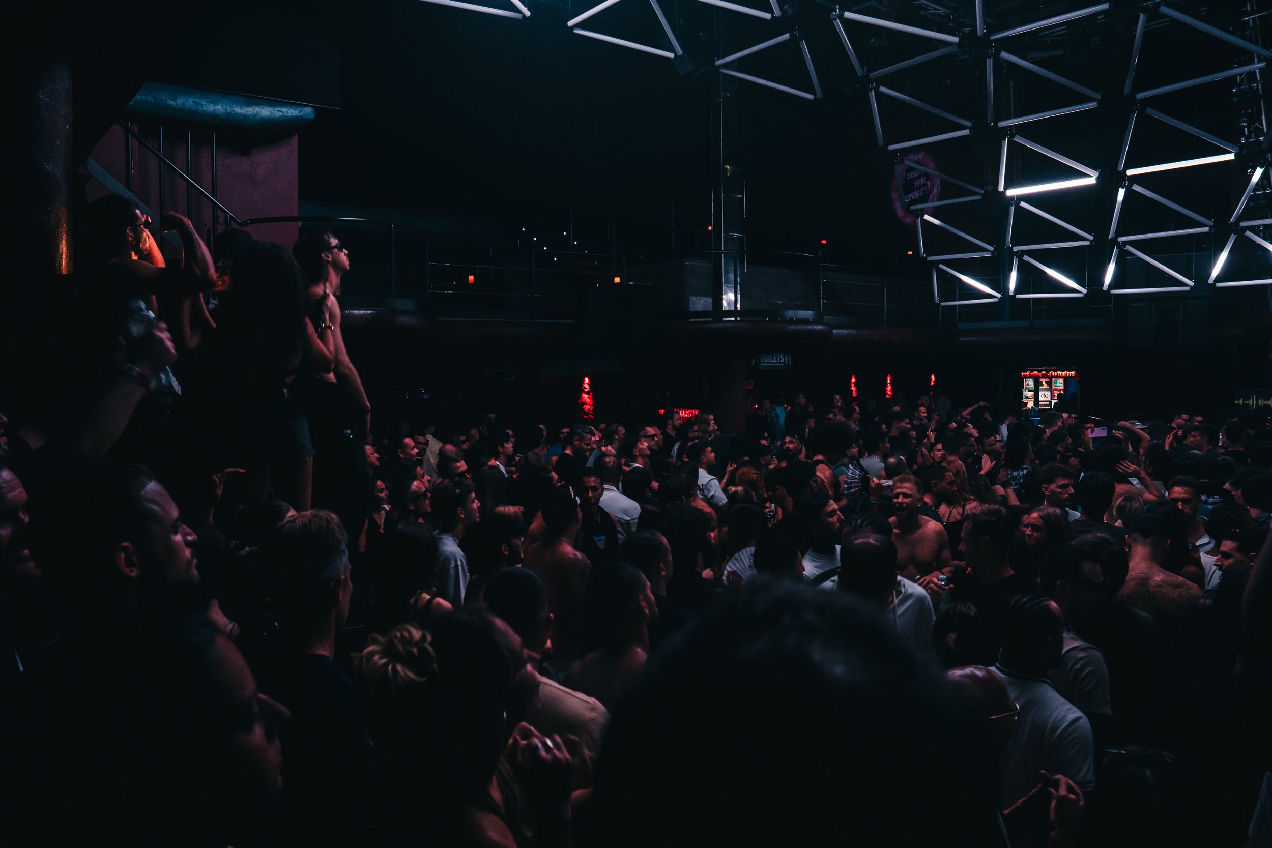 A large crowd of people gathered in a dark indoor venue with modern lighting structures on the ceiling, some people standing and others sitting, attending an event or concert.
