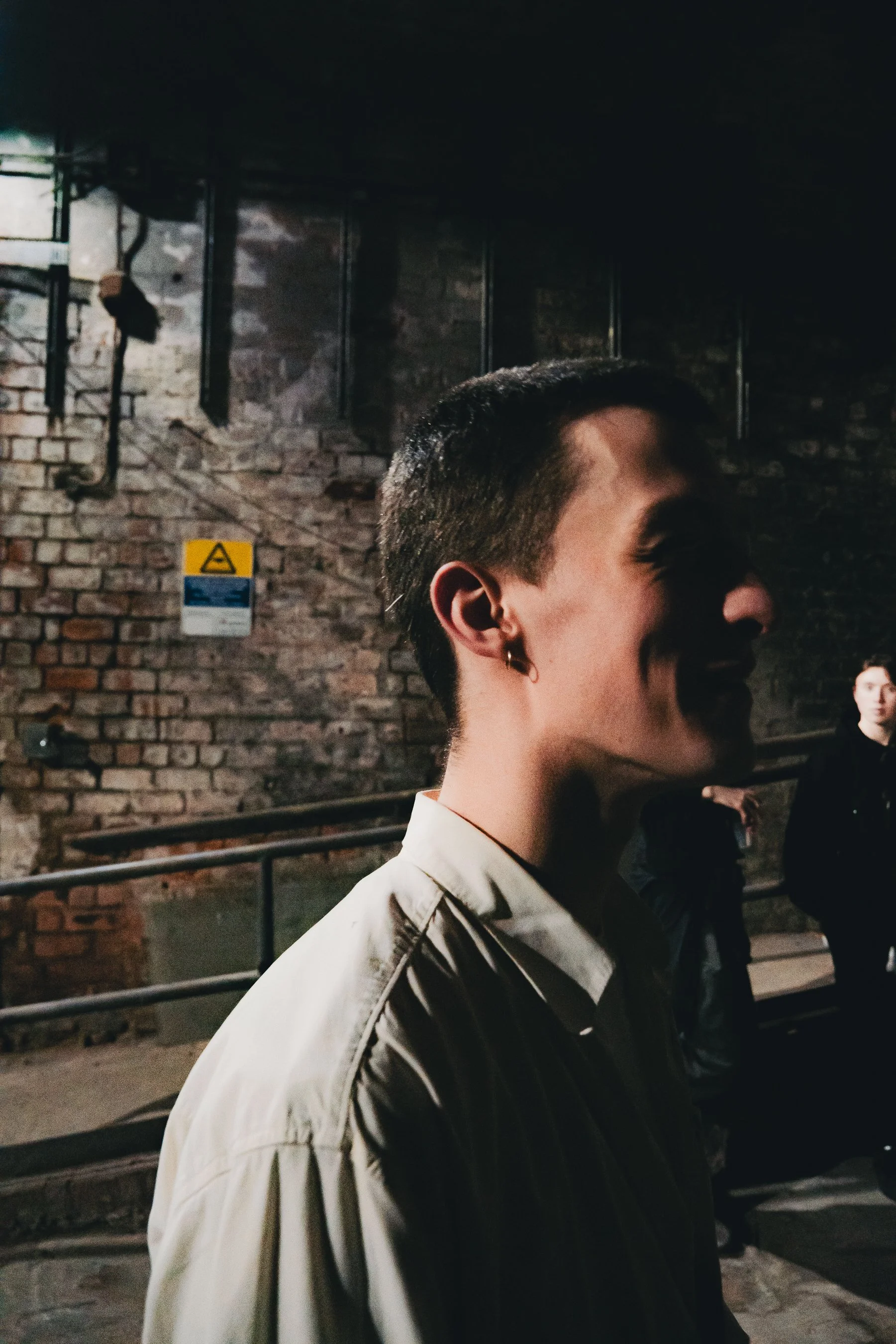 A person with short dark hair and earrings, wearing a light-colored jacket, standing in front of a brick wall at night in an urban setting.