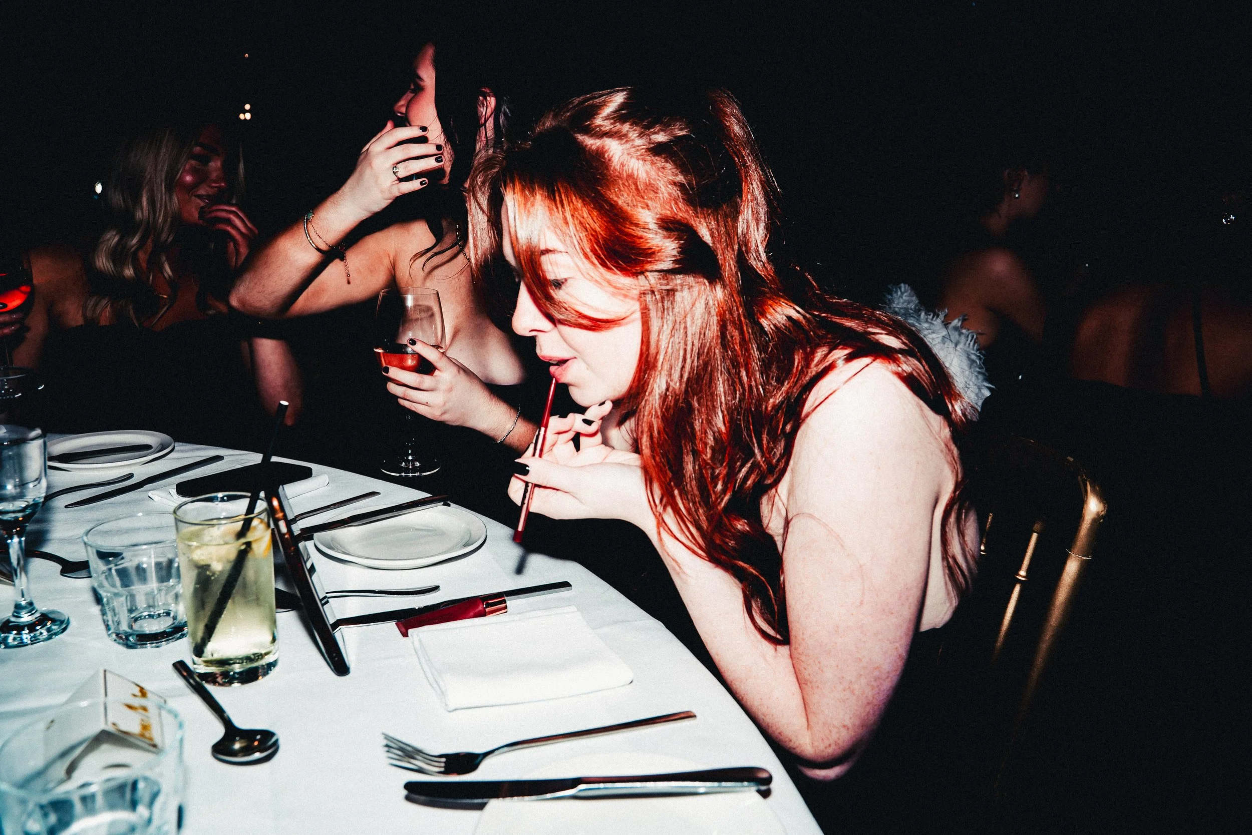 Five women sitting at a dinner table, engaging in conversation and drinking wine, in a dimly lit setting.