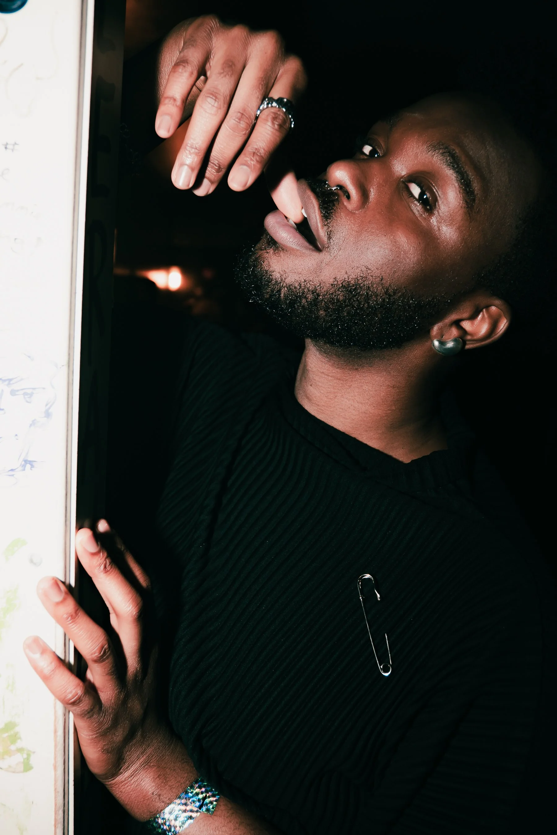 Close-up of a person with dark skin, a beard, and a mustache, wearing a black ribbed shirt, silver earring, and a silver bracelet, holding a phone with a polka dot case near their face, with a dark background.