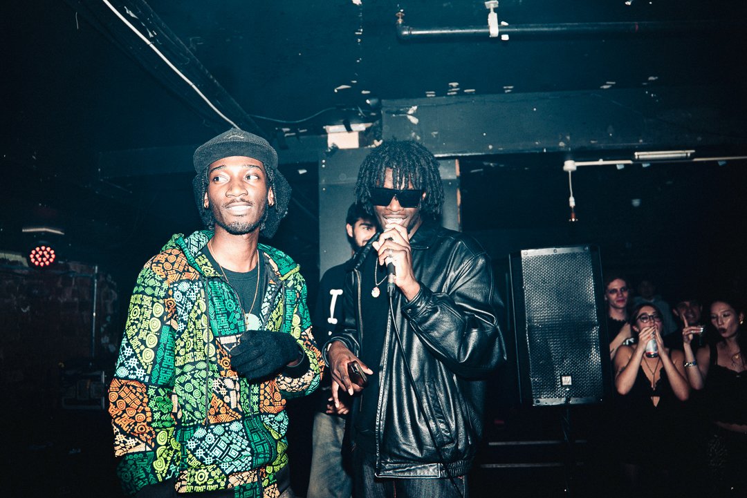 Two men are on stage at a concert or event. One is wearing a colorful patterned hoodie, a gray beanie, and black gloves. The other is wearing sunglasses, a black leather jacket, and has dreadlocks, holding a microphone. Several people are visible in 