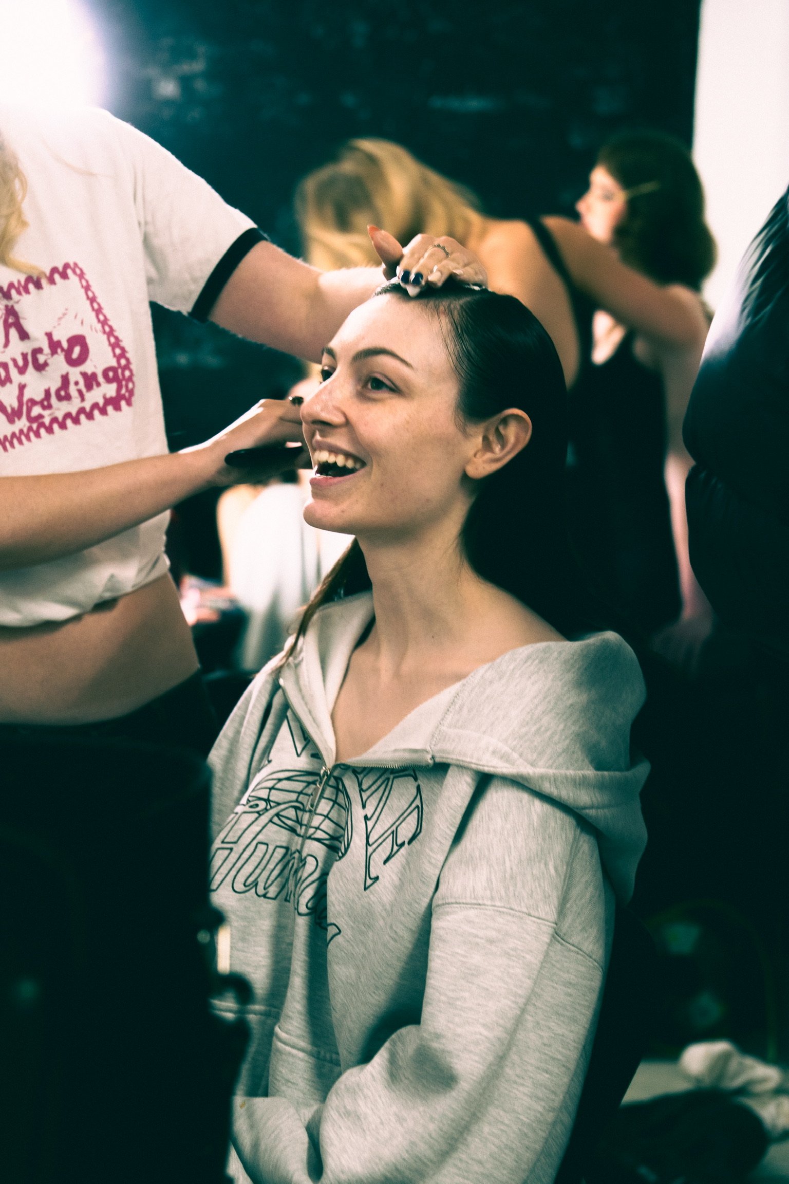 A young woman with dark hair sitting in a chair, smiling as she gets her hair styled, with two women in the background also styling hair.