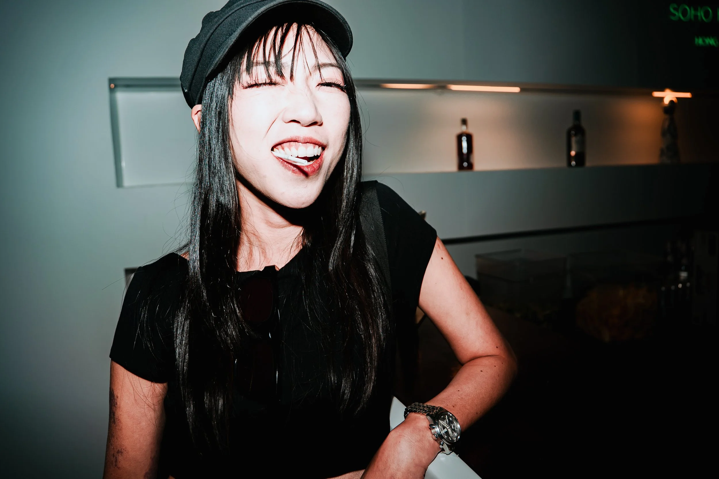 A woman with long black hair, wearing a black top, a black hat, and a silver watch, smiling and showing her teeth in a lively setting with a blurred background featuring bottles on a shelf and a neon sign.