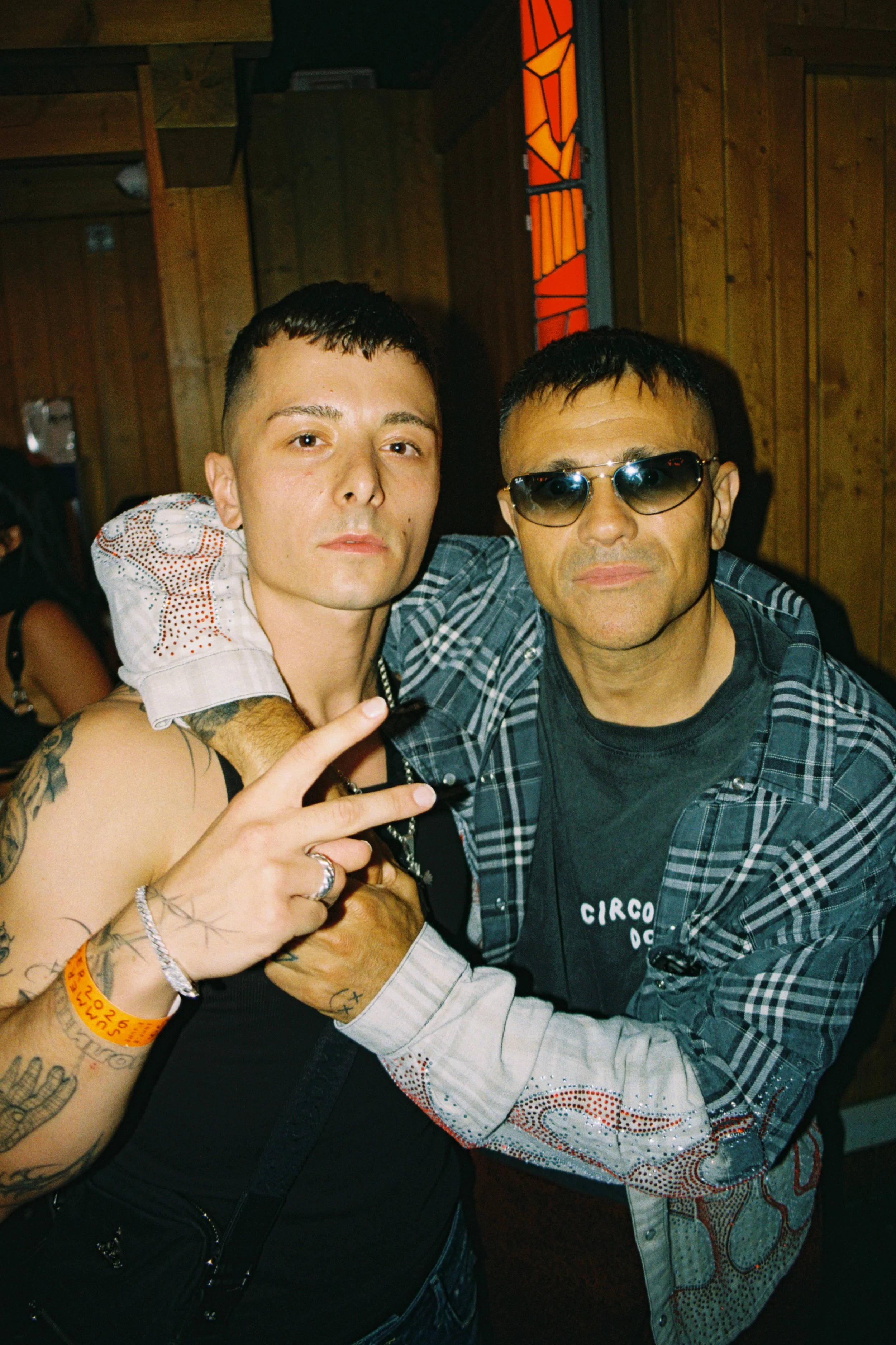 Two men are posing closely together, one wearing sunglasses and a plaid shirt, the other with tattoos and a black top, inside a wooden-paneled room with colored stained glass in the background.