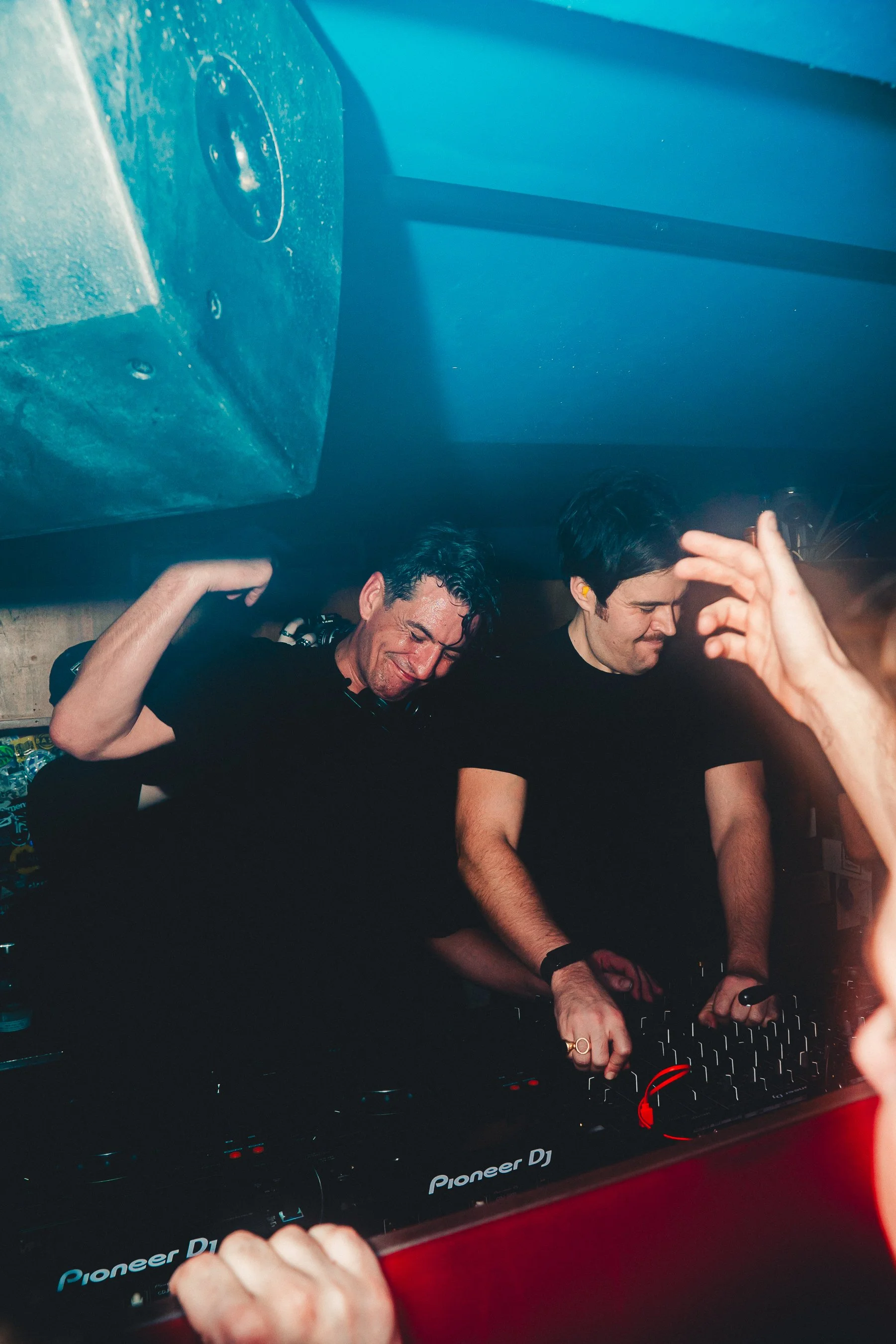 Two young males DJing at a nightclub or party, with one wearing yellow earplugs and the other wearing glasses and headphones, with a blue wall and DJ equipment in the background.