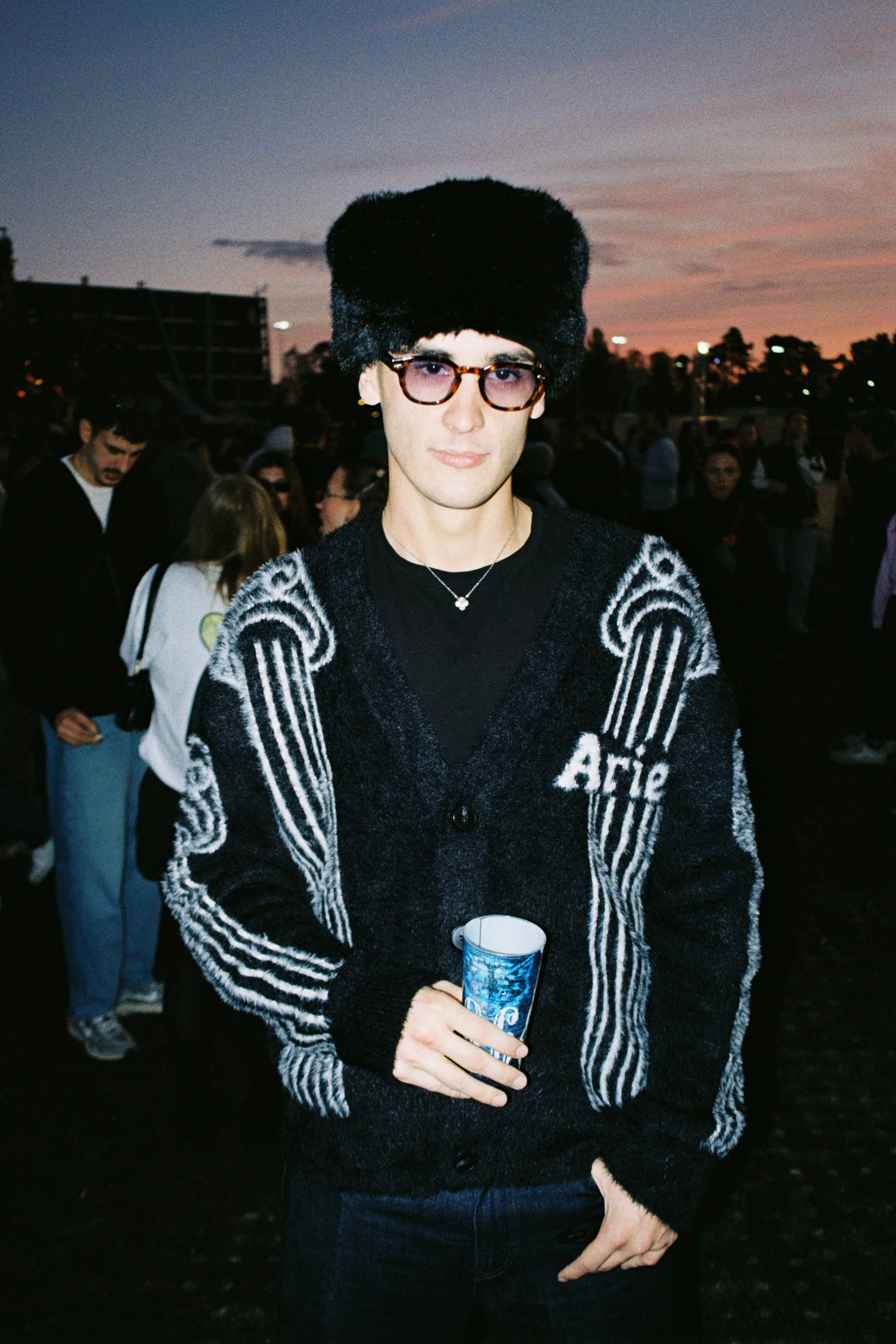 Young man wearing a large black fur hat, patterned black and white sweater, and glasses, holding a blue beverage cup, at dusk with a crowd and sunset sky in the background.