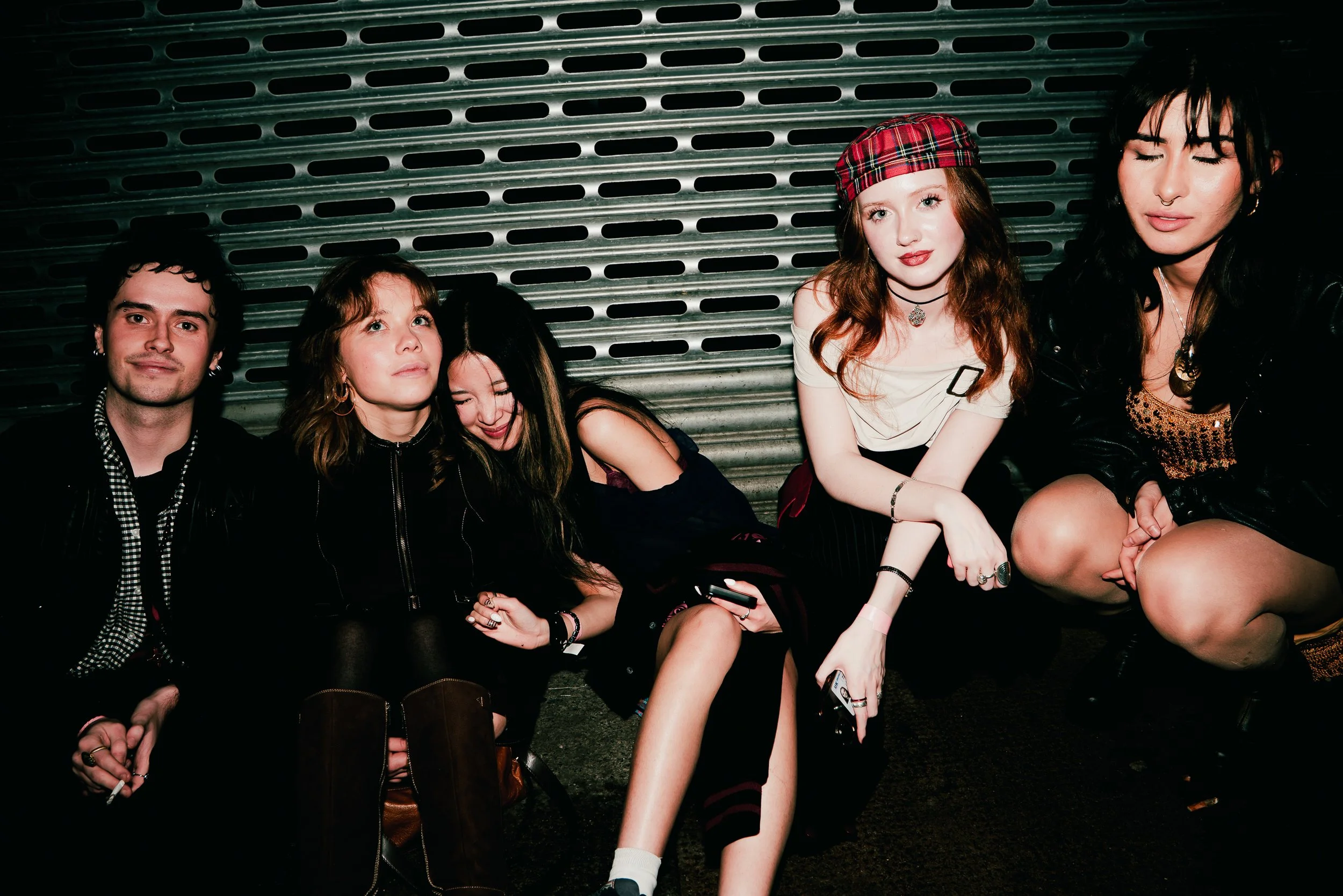 A group of five young people sitting against a metal roll-up door, with diverse hair colors and styles, dressed in casual and alternative fashion, some with makeup and jewelry, posing for a photo.