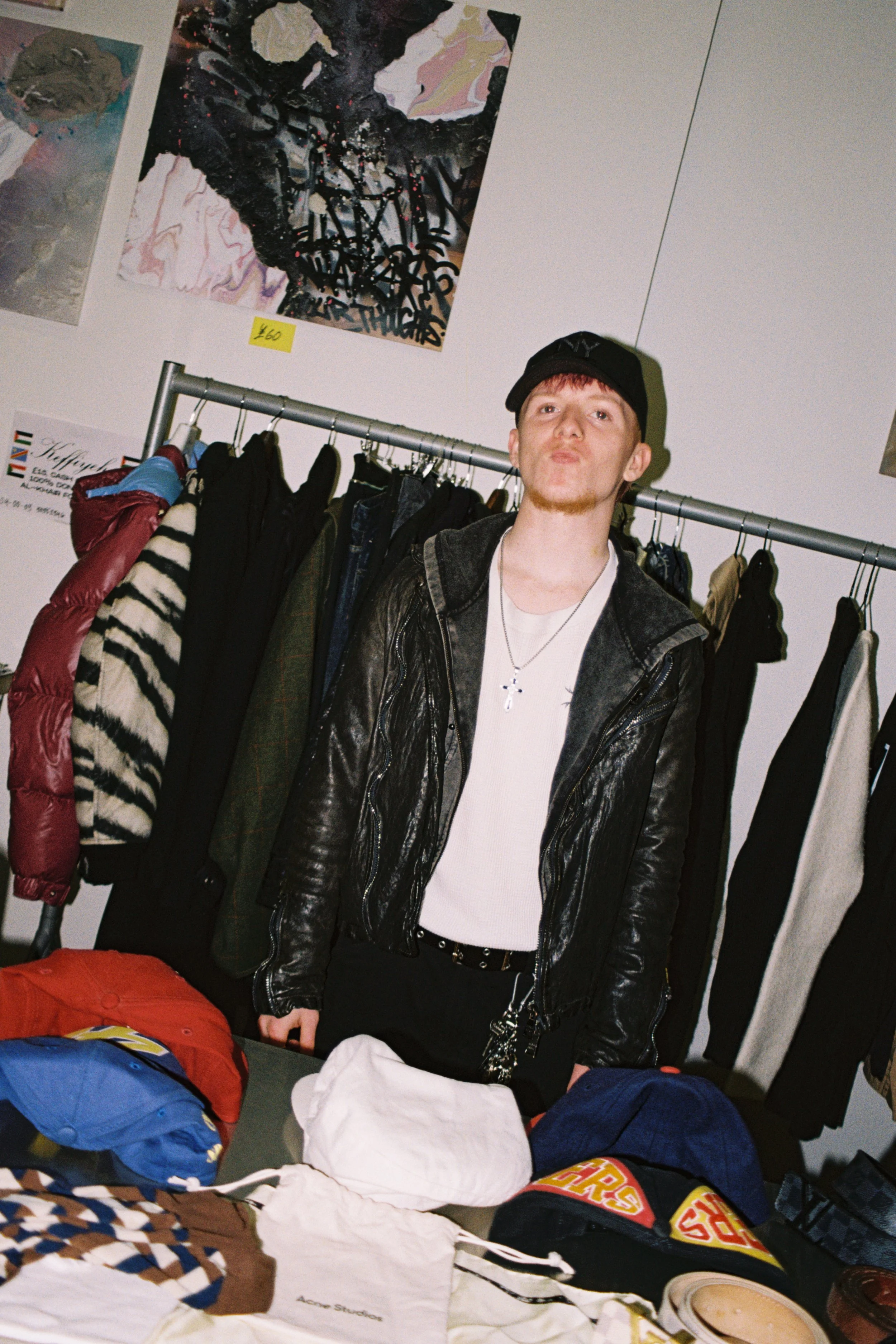 A young man with red hair, wearing a black cap, leather jacket, and a white t-shirt with a silver cross necklace, standing in front of a rack of jackets and coats. On the table in front of him are piles of clothes and bags, with colorful clothing ite