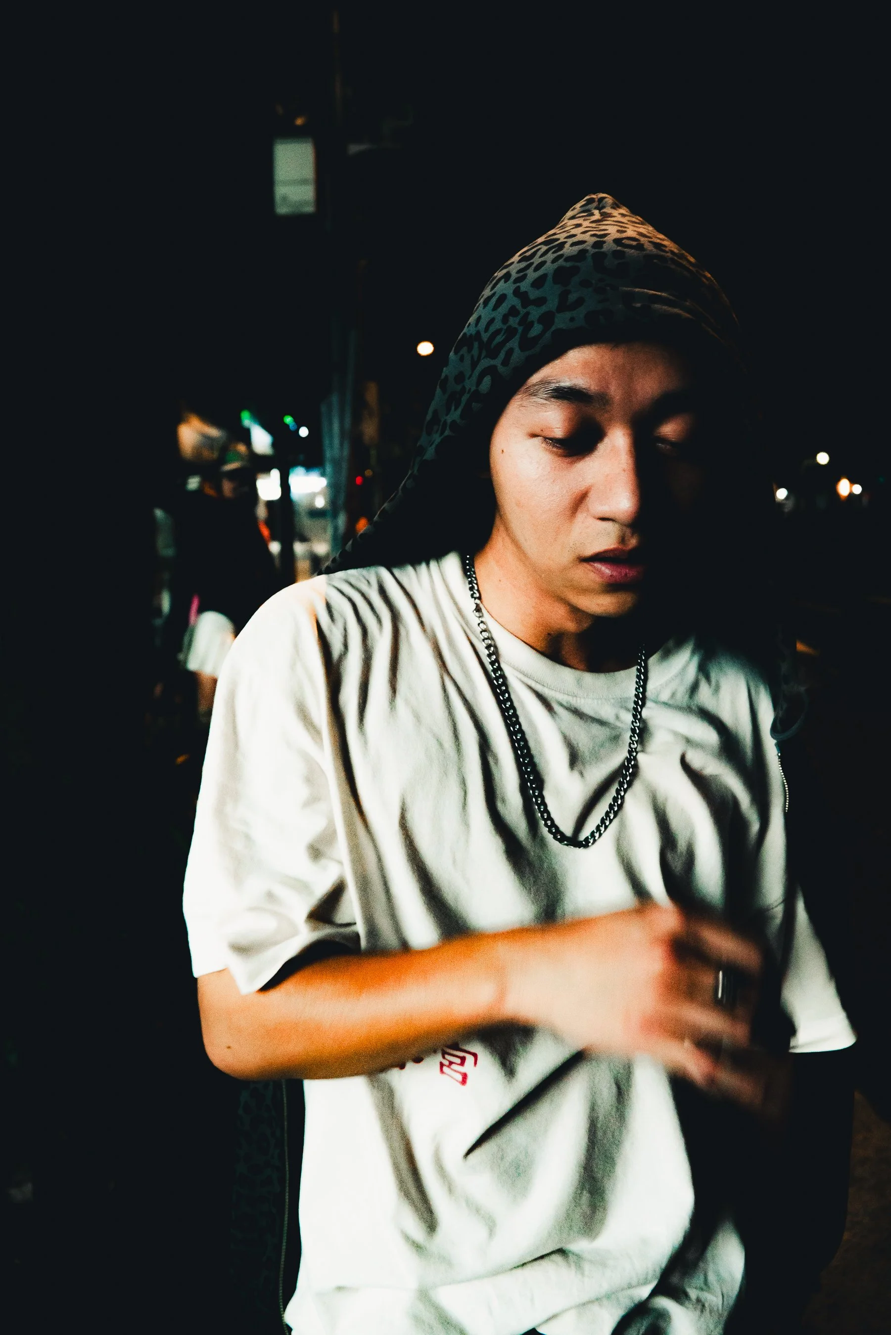 A young man with a leopard print hood covering his head, wearing a white t-shirt and chains, standing in a dark, urban outdoor setting at night with blurred city lights in the background.