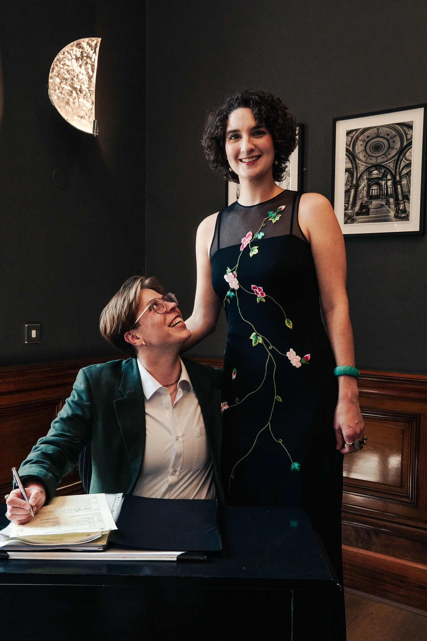 A woman in a black dress with floral embroidery smiling and standing next to a seated person wearing glasses and a dark green blazer, signing papers on a black table in a room with dark walls, framed artwork, and a moon-shaped light fixture.
