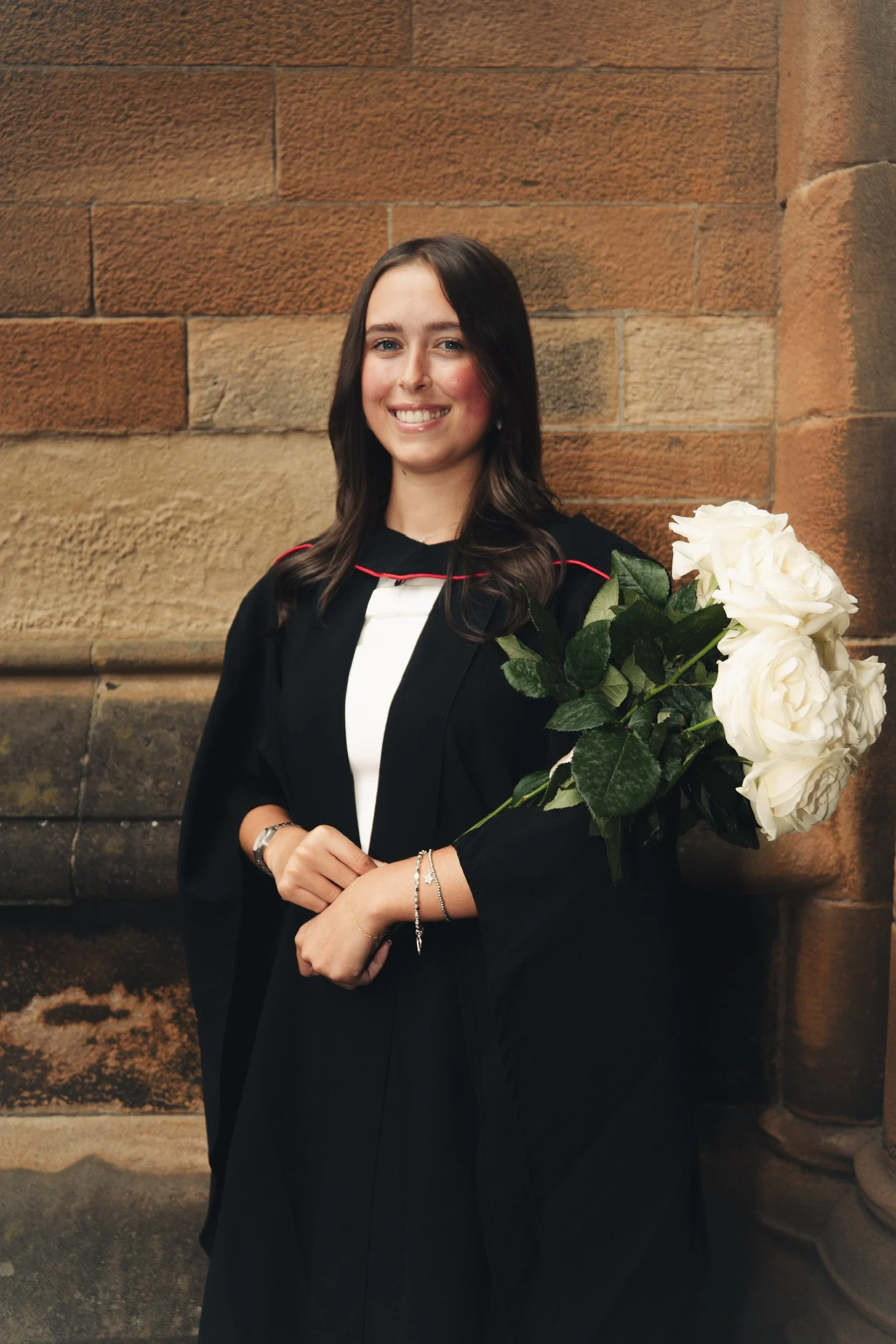 A young woman in a graduation gown and cap holding white roses, smiling, standing against a brick wall.