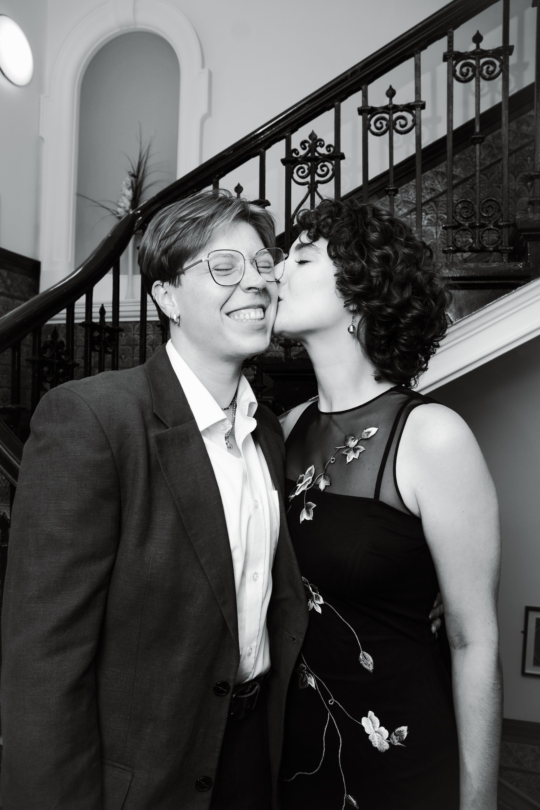Two women, one with short hair and glasses and the other with curly hair, are indoors. The woman with glasses is smiling as the curly-haired woman kisses her cheek. They are standing near a staircase with ornate railing, and there is a decorative flo