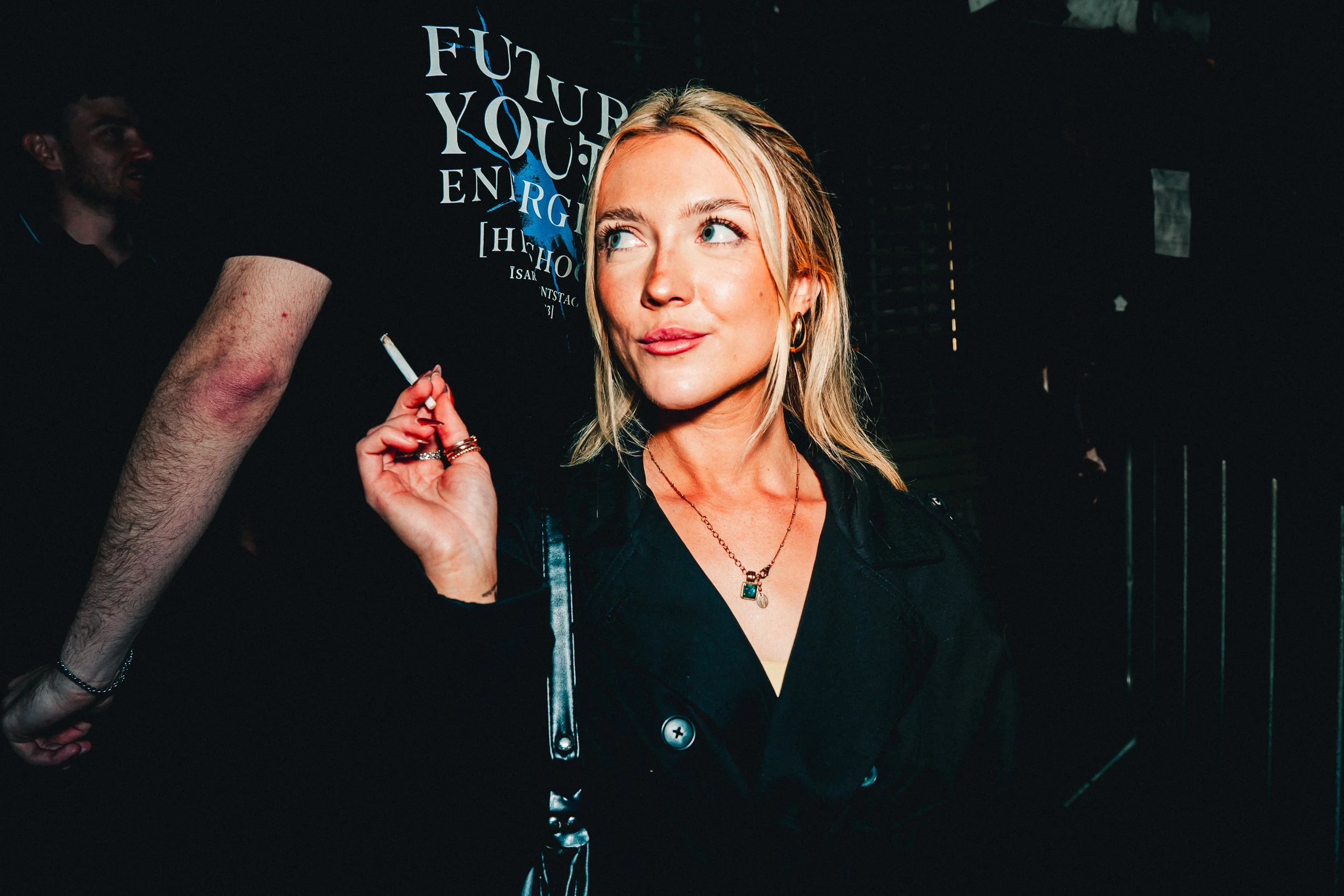 A woman with blonde hair and blue eyes holding a cigarette, standing in front of a black wall with white and blue lettering that says 'FUTURE YOU' and other words.