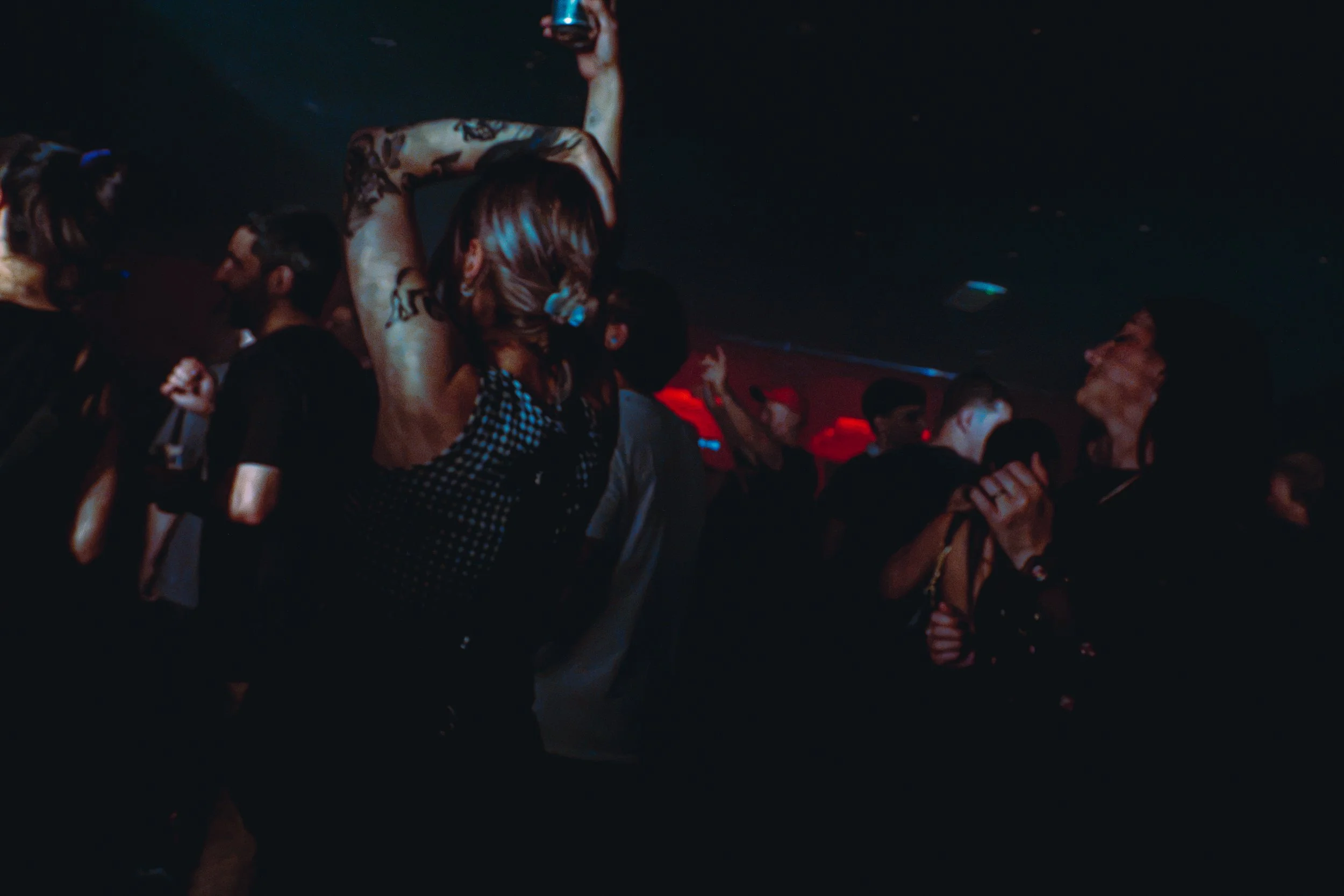 People dancing in a dark nightclub with colorful lighting, some embracing or holding drinks.