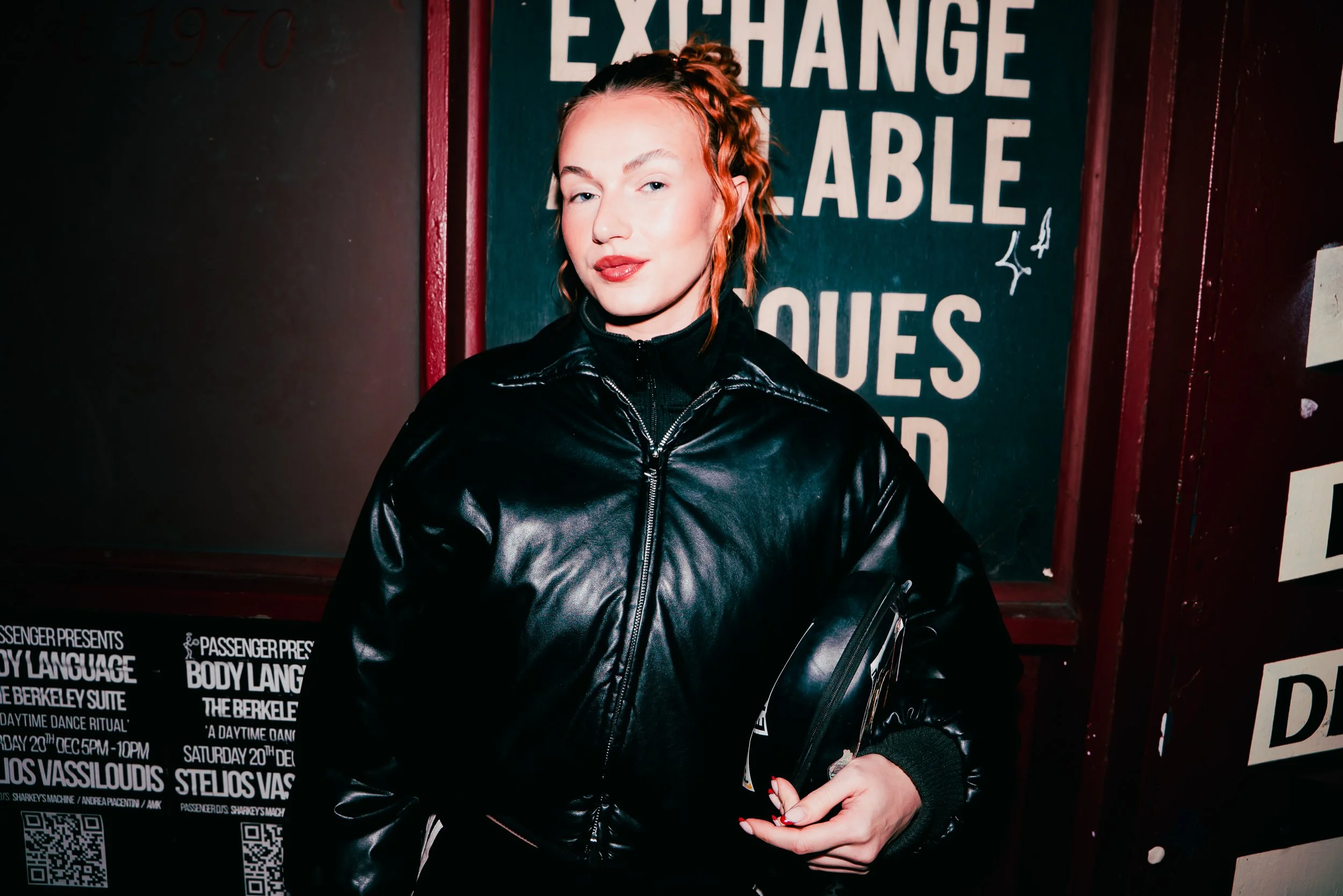 A woman with curly red hair and pale skin poses in front of a sign that says 'Exchangeable'. She is wearing a shiny black jacket with a black turtleneck underneath. Her eyes are slightly squinted, and she has a confident expression with red lipstick.