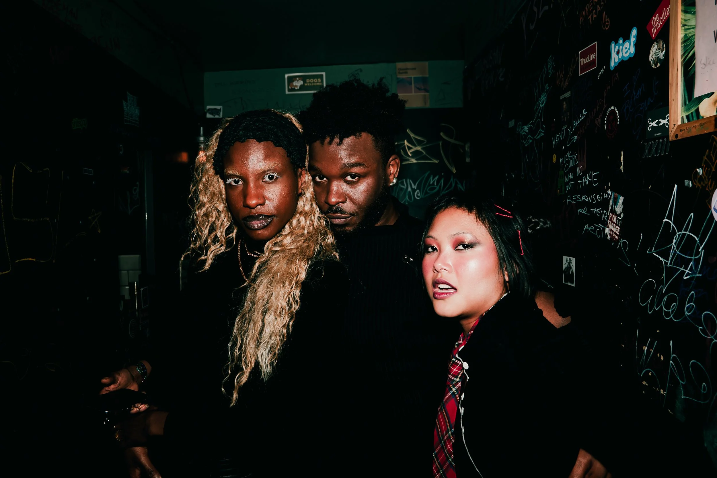Three young women with distinctive hairstyles and bold makeup pose together in a dimly lit room with graffiti-covered walls.