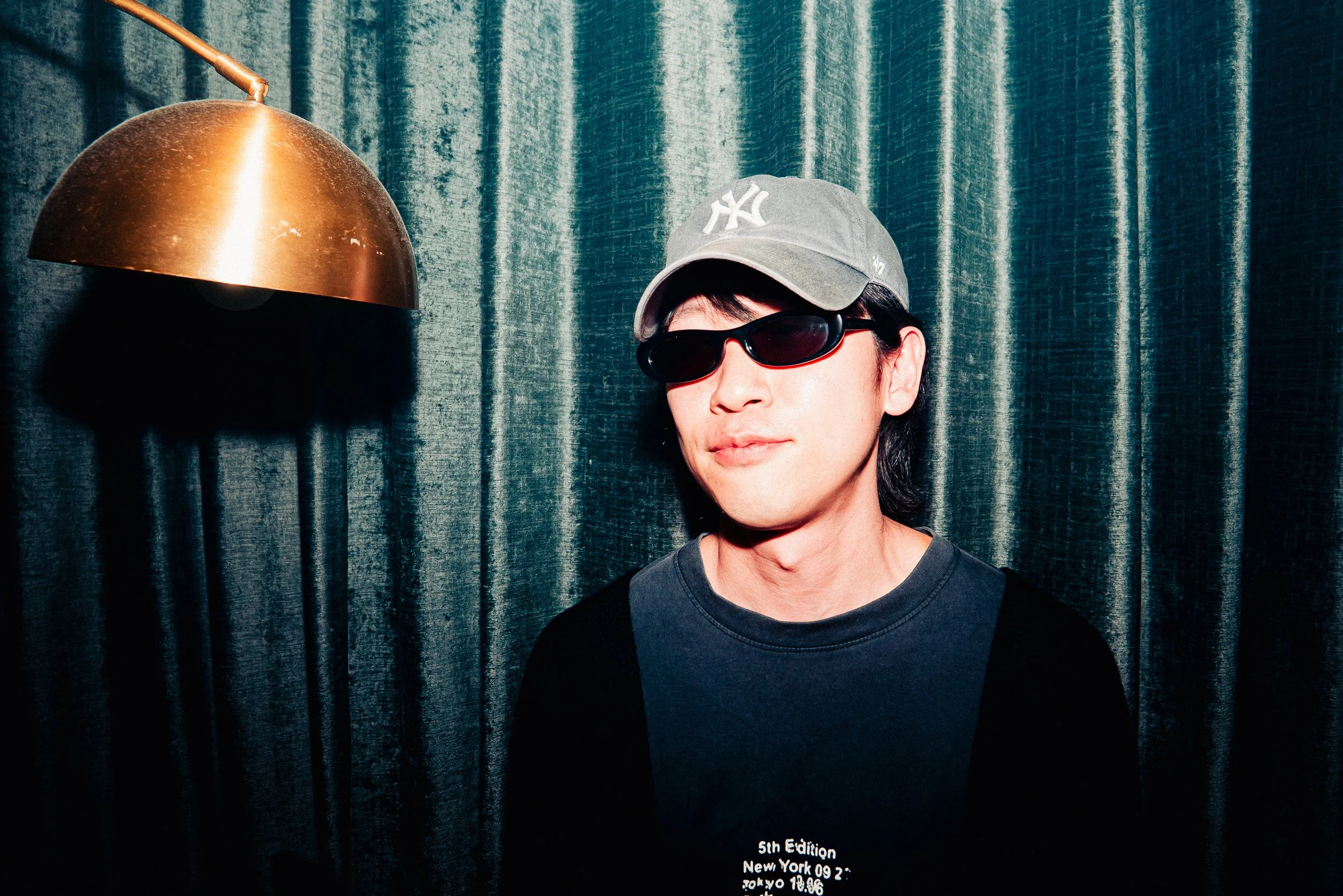 A person wearing a gray New York Yankees cap and sunglasses poses in front of dark teal curtain with a gold-colored lamp on the left. There is some white text at the bottom right of the image.