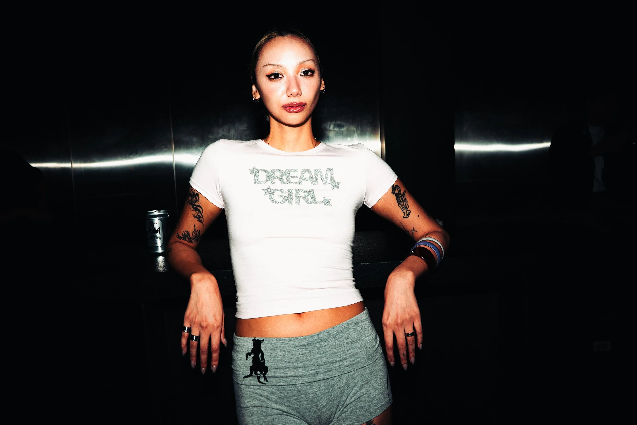 A woman with tattoos and hoop earrings wearing a white t-shirt that says 'DREAM GIRL' and gray shorts with a black dog silhouette, standing against a dark background.