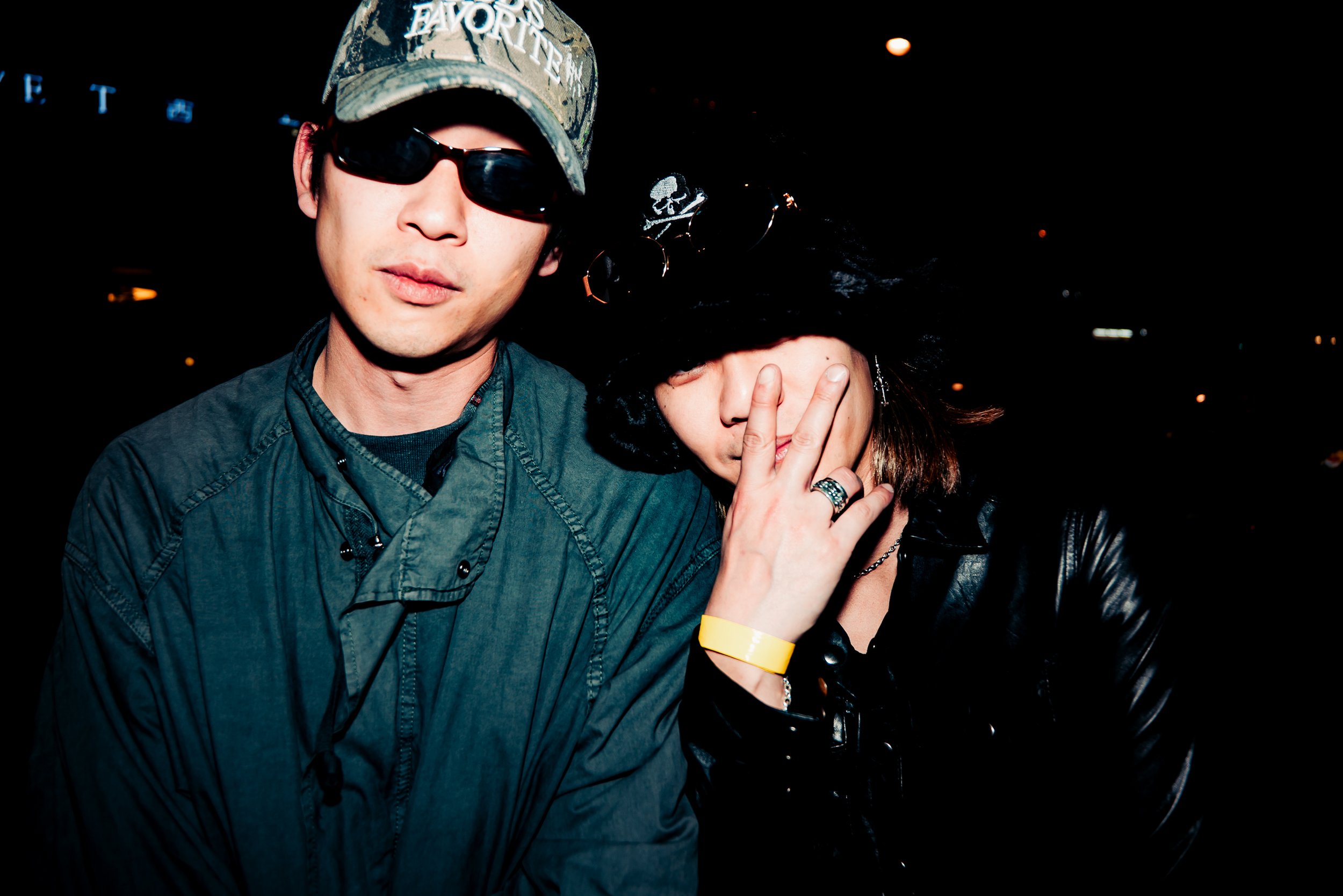 A man wearing a camouflage baseball cap with the words "FAVORITE" and dark sunglasses, and a woman with a black curly wig and black leather jacket, covering her face with her hand.