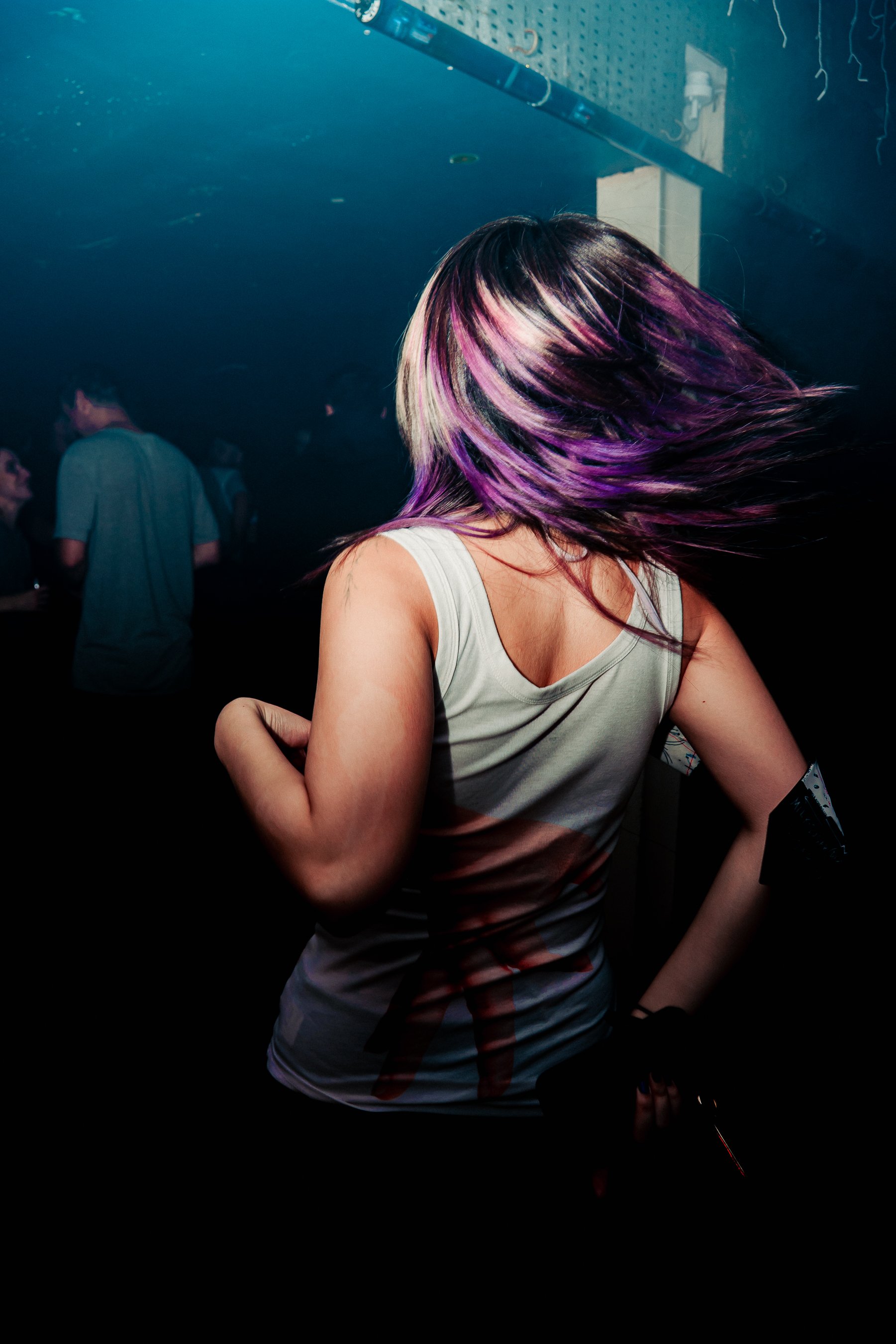 A woman with black and purple hair dancing in a dark nightclub with other people in the background.