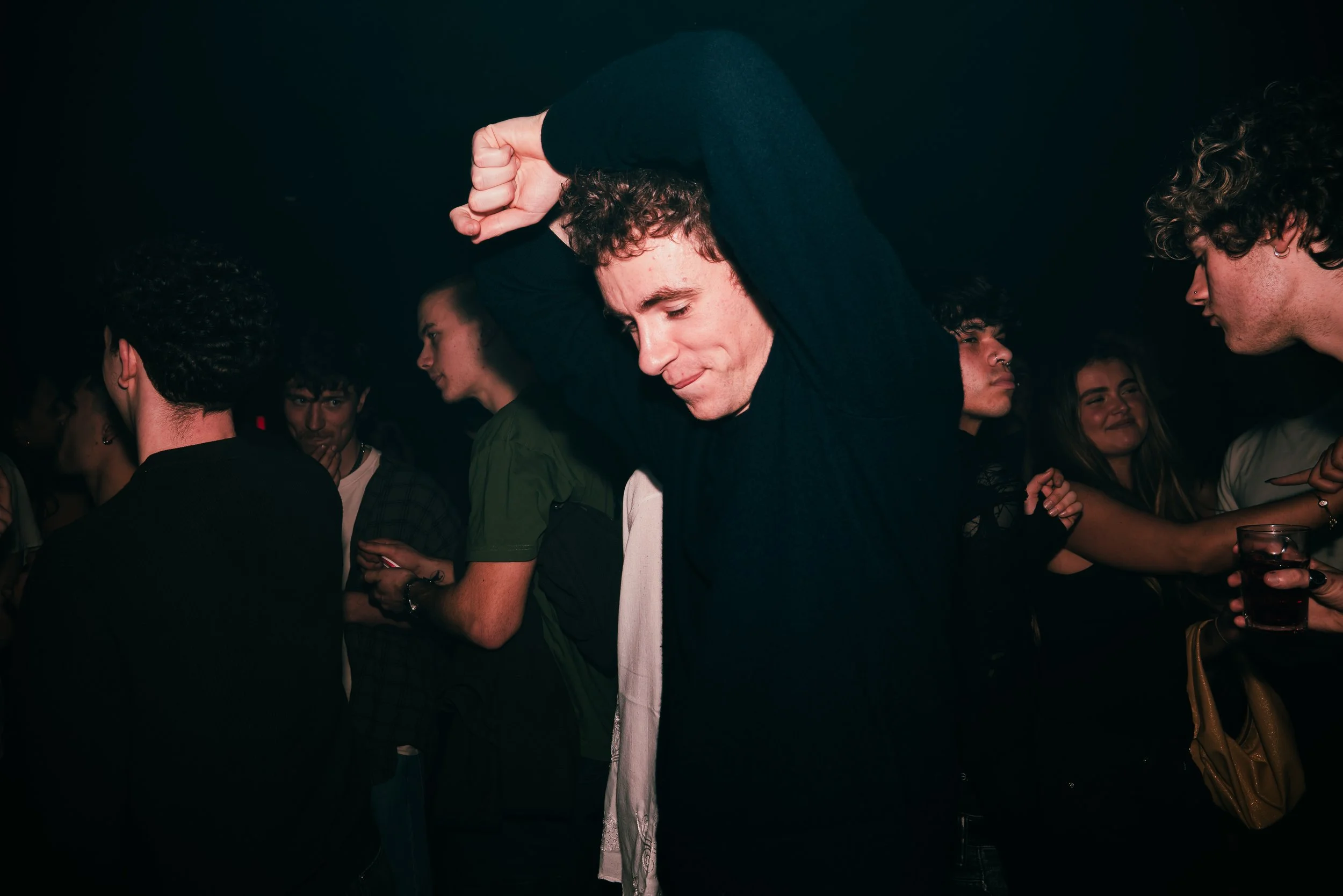 A group of young people socializing at a party or club, with one person in the foreground dancing and others talking in the background.