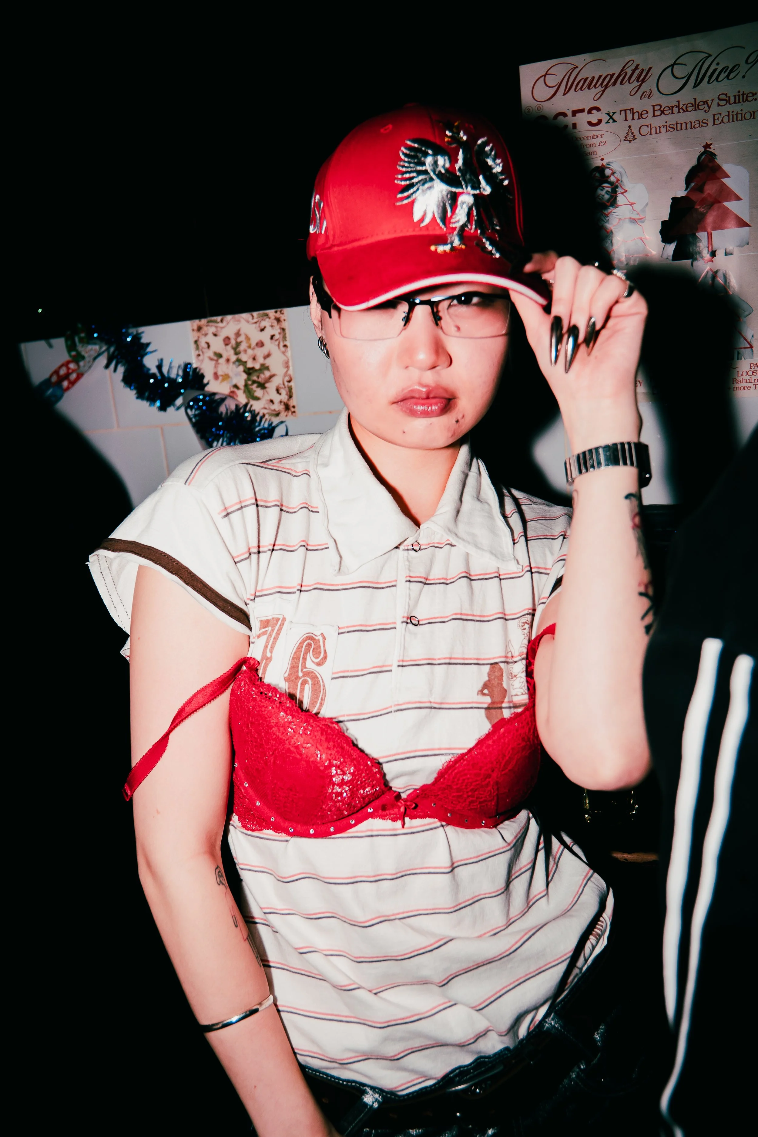 A woman wearing a red baseball cap with a pegasus design, glasses, a striped white and gray short-sleeve shirt, and a red sequined bra, is adjusting her cap while standing indoors at a decorated event with Christmas decorations and a sign in the back