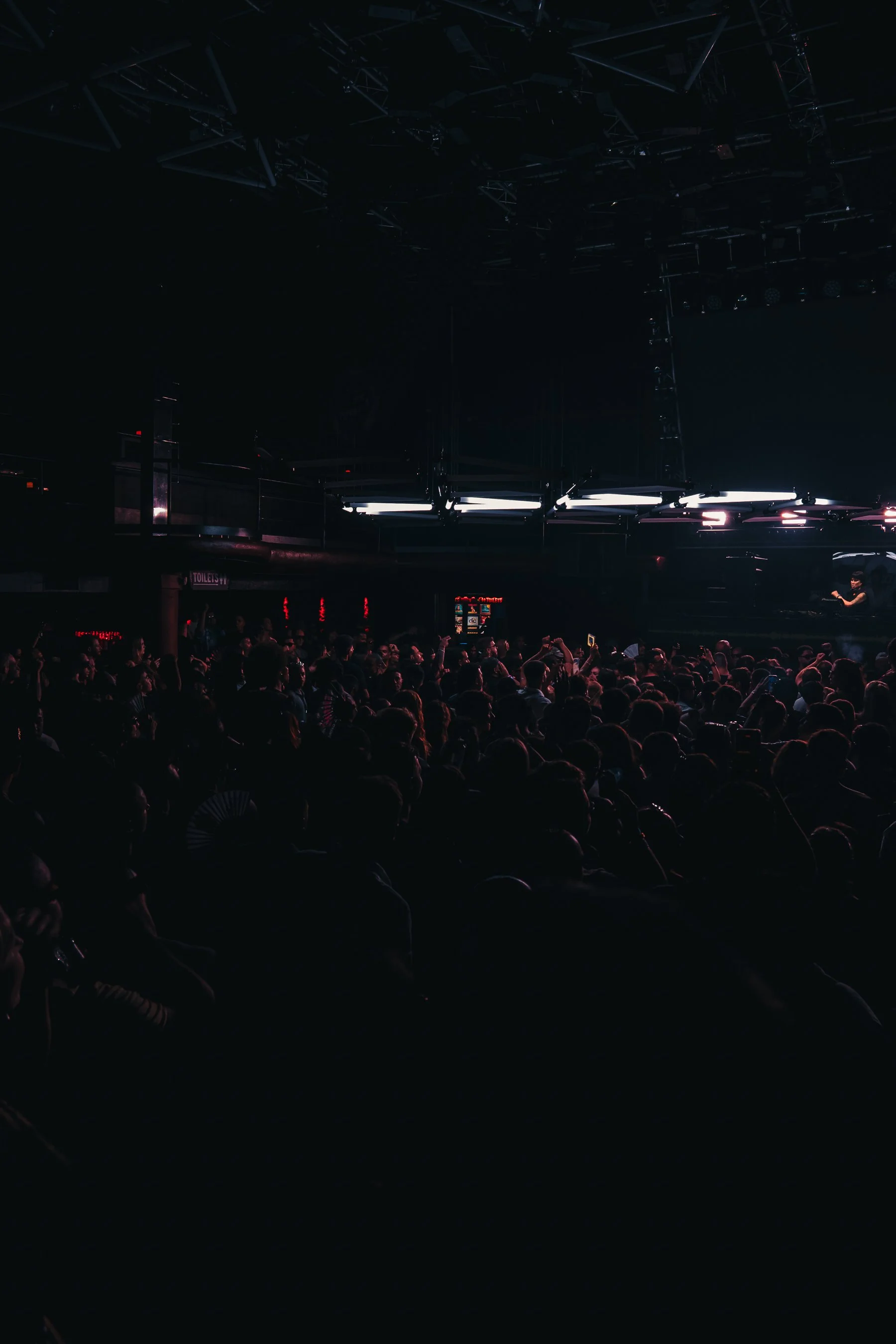 Audience seated in dark concert venue with stage lights and lighting fixtures hanging above.